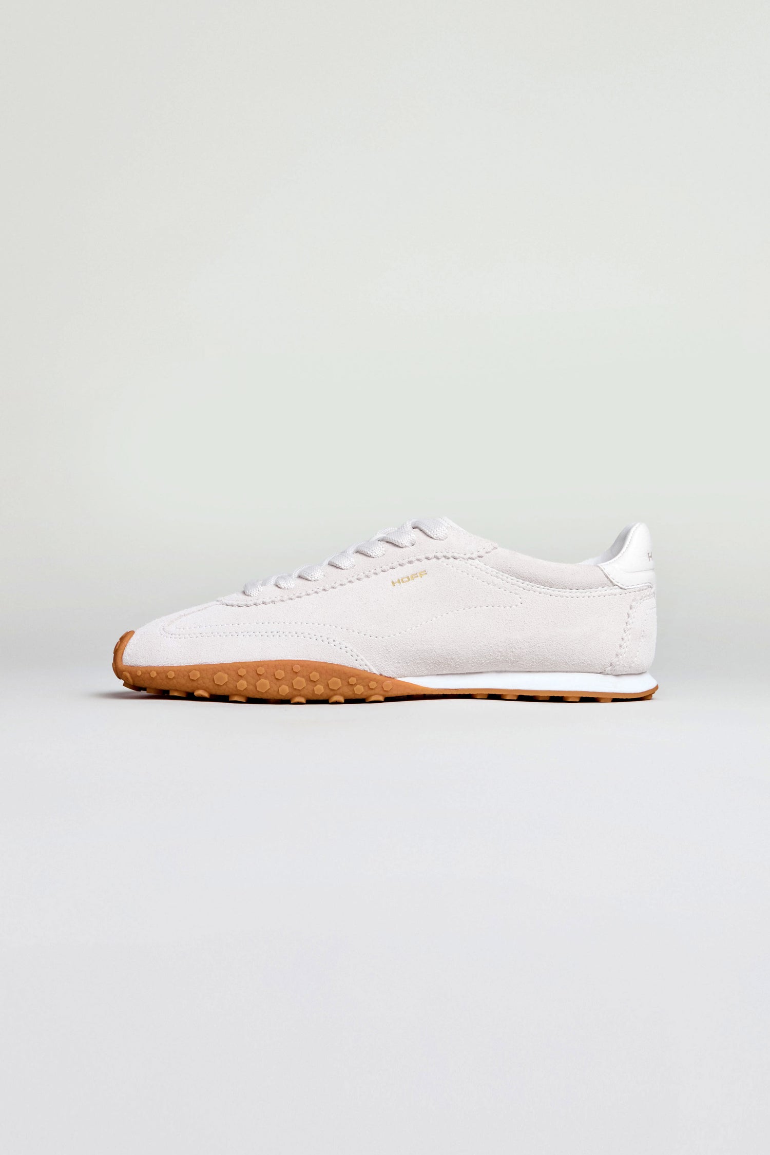 The Bridge Light Grey Suede Trainers, shown in profile against a plain background, feature a white low-top design with a gum rubber sole and subtle suede accents inspired by retro running shoes.