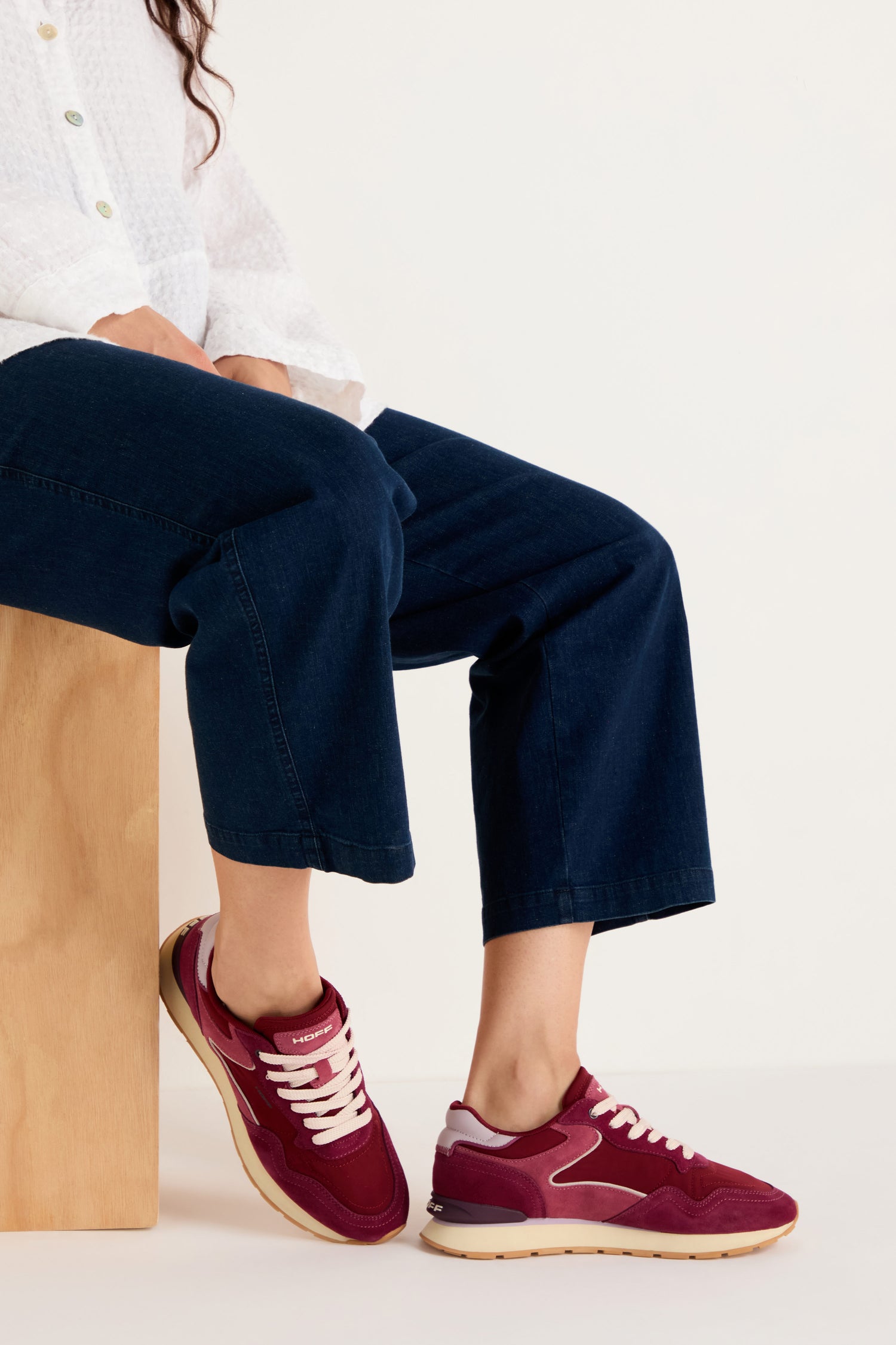 A person seated on a wooden block wears a white shirt, dark blue jeans, and City Trainers—premium suede footwear with white laces—against a plain light background.
