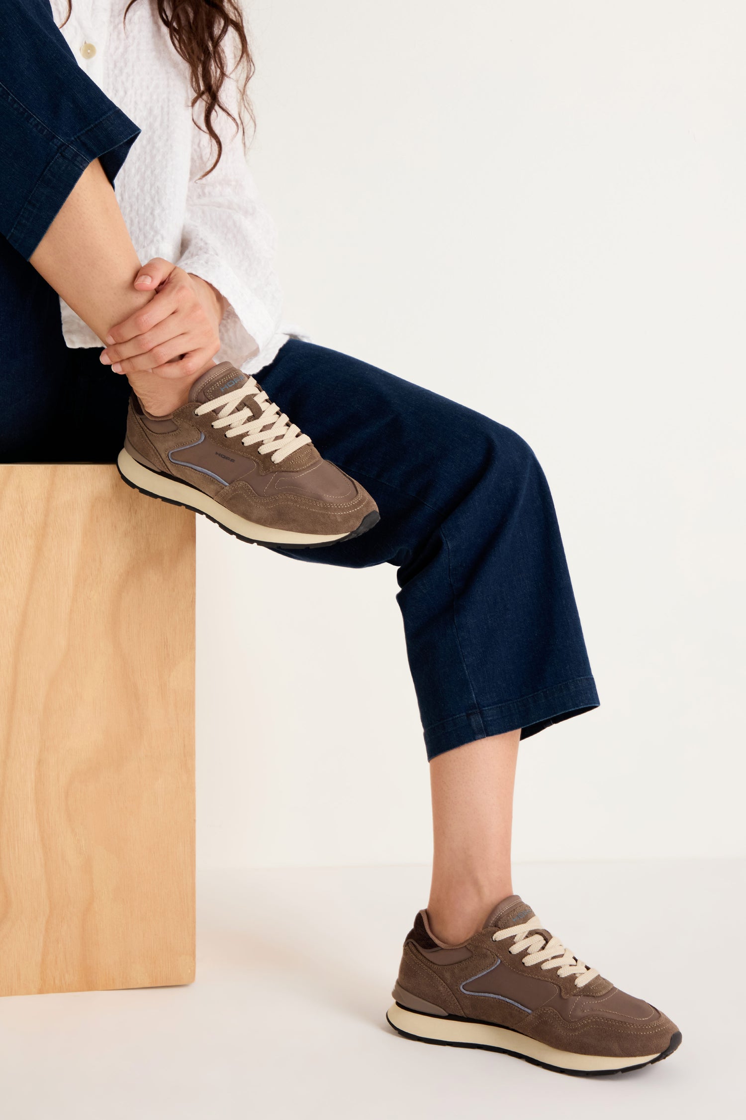 A person wearing dark blue pants and City Trainers with premium suede sits on a wooden block against a plain white background.