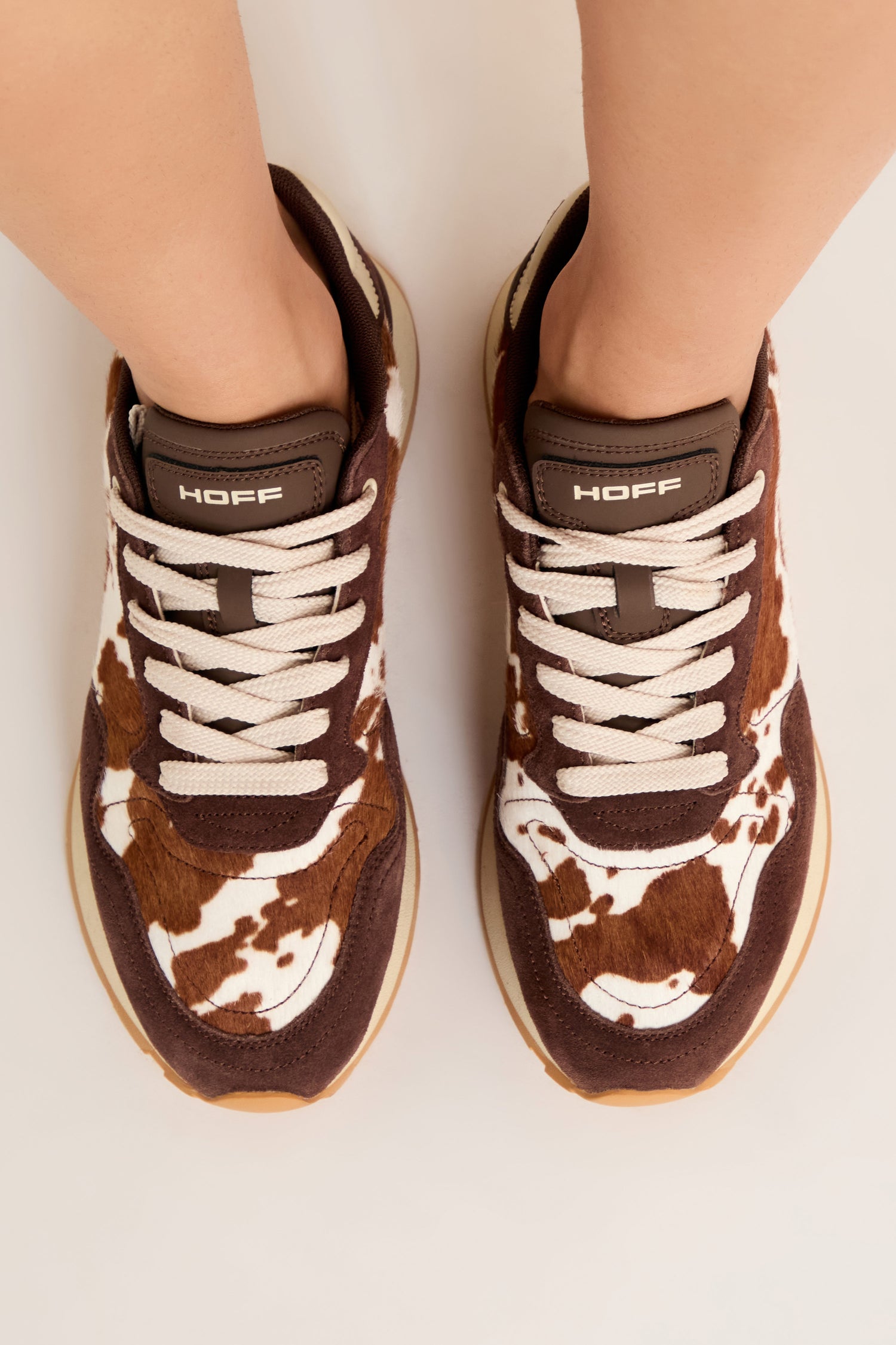 A person wears Animal Print City Trainers featuring a brown and white cowhide pattern, premium suede accents, and white laces, photographed from above against a plain background.