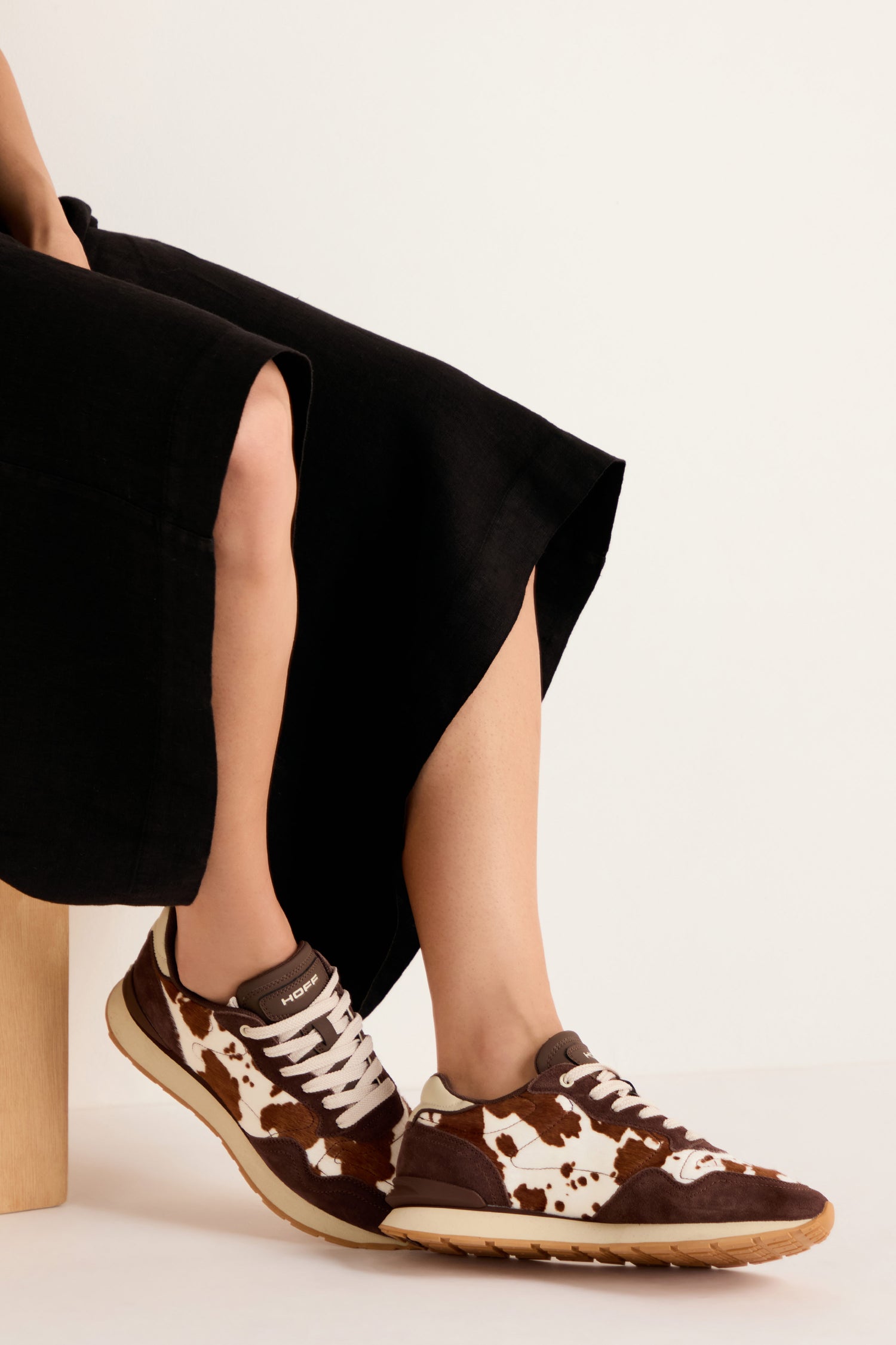 A person wearing a long black skirt sits on a wooden block, showcasing the Animal Print City Trainers—premium suede sneakers featuring a brown and white cowhide pattern with beige soles—against a plain background.