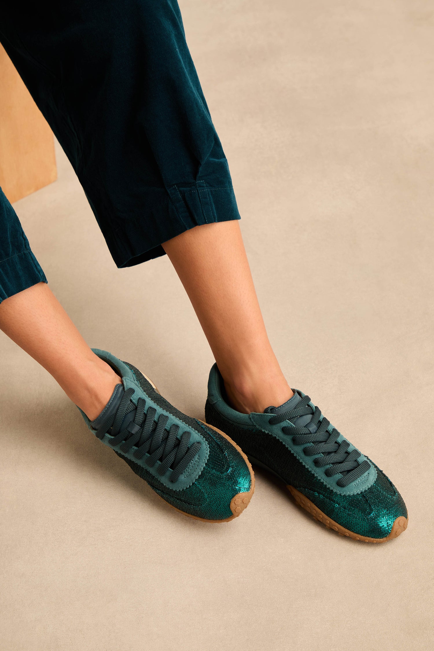Person wearing teal corduroy pants and stylish Sequin Bridge Trainers—teal suede sneakers with brown soles and a comfortable memory-foam insole—standing on a beige floor.
