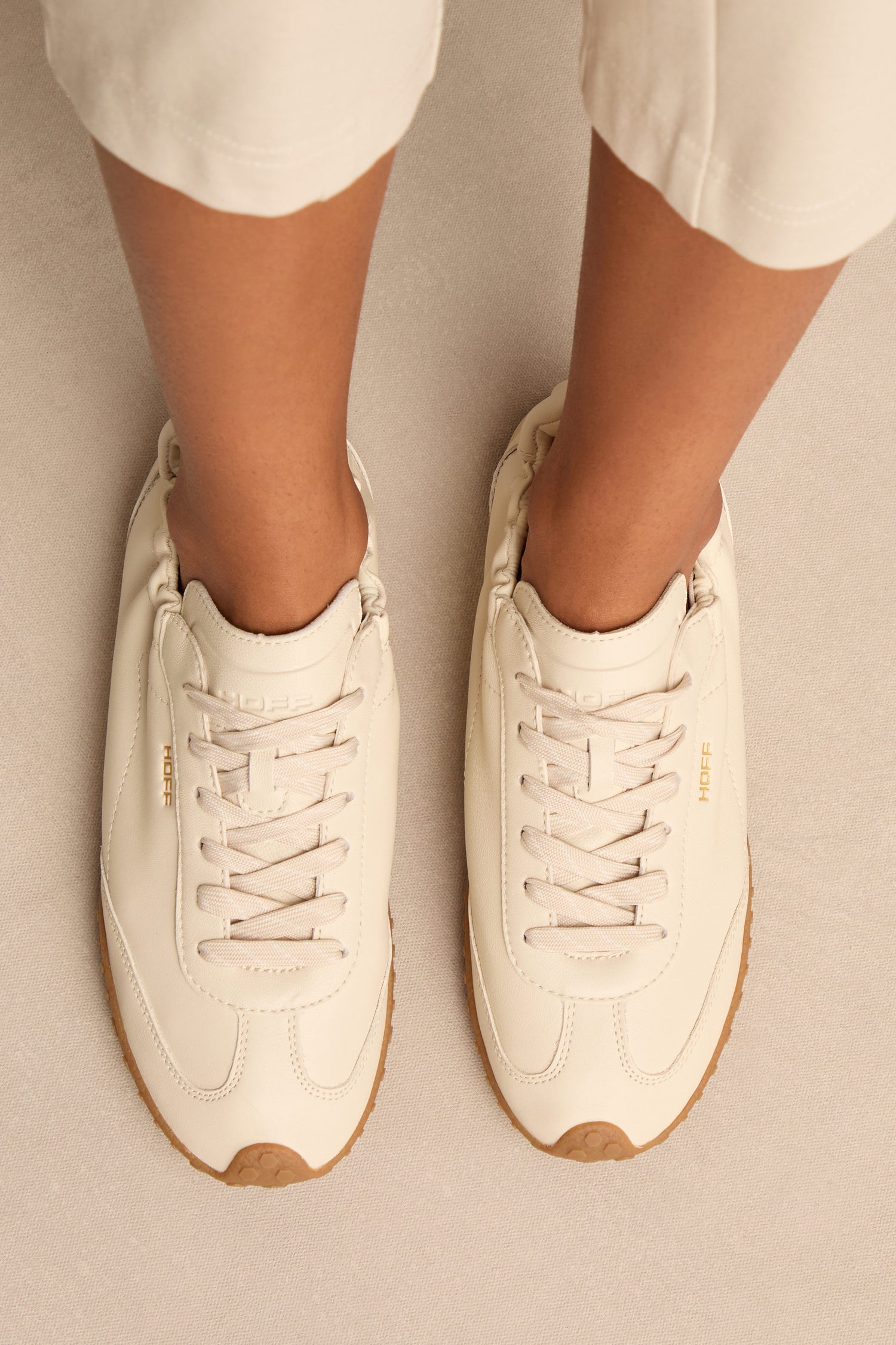 Person wearing Napa Leather Bridge Trainers—off-white, low profile shoes with tan soles and beige laces—standing on a light-colored surface. Only lower legs and feet are visible.