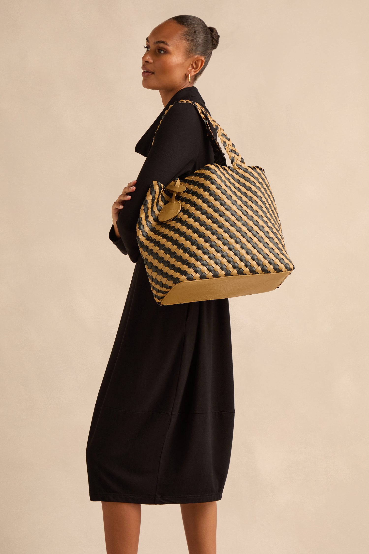 A woman in a black dress stands sideways, carrying the Reversible Braided Shopper in bold black and tan hues over her shoulder against a neutral background.