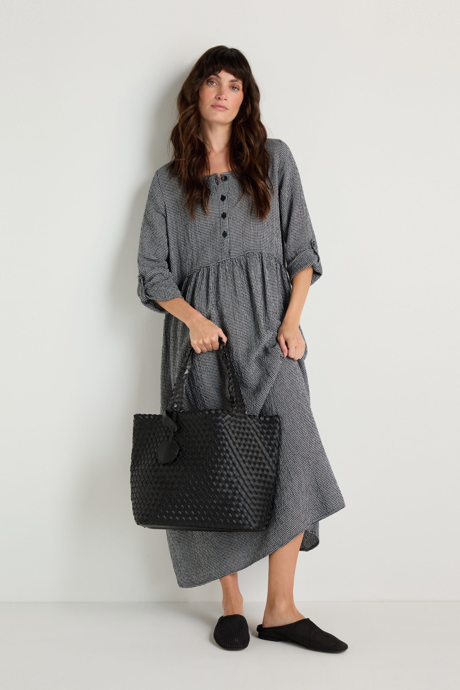 A woman in a loose gray long-sleeve dress stands against a white wall, holding a Woven Reversible Bag and wearing black slip-on shoes.