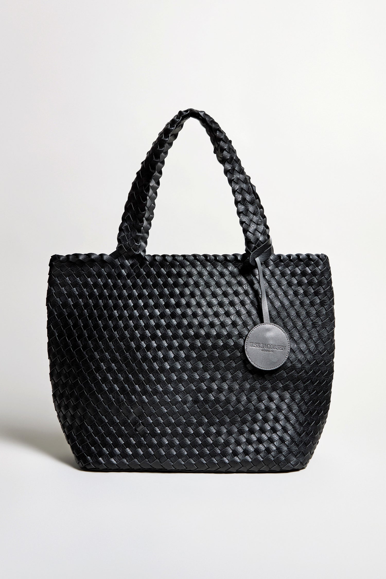 The Woven Reversible Bag features a black basket weave design, two handles, and a round tag, shown against a plain light background.