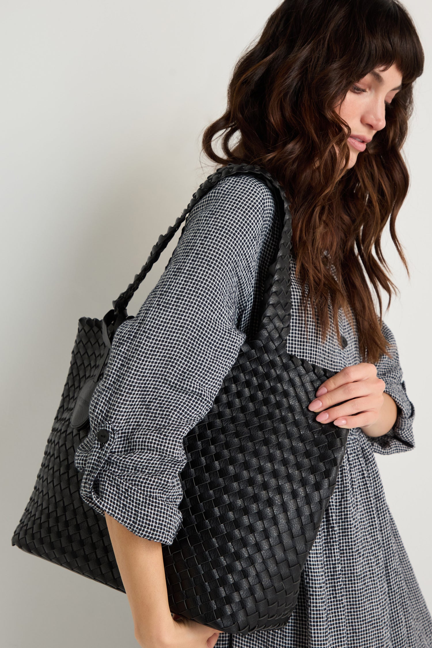 A woman in a checkered dress stands against a plain light background, carrying the Woven Reversible Bag—a large black vegan leather tote—over her shoulder.