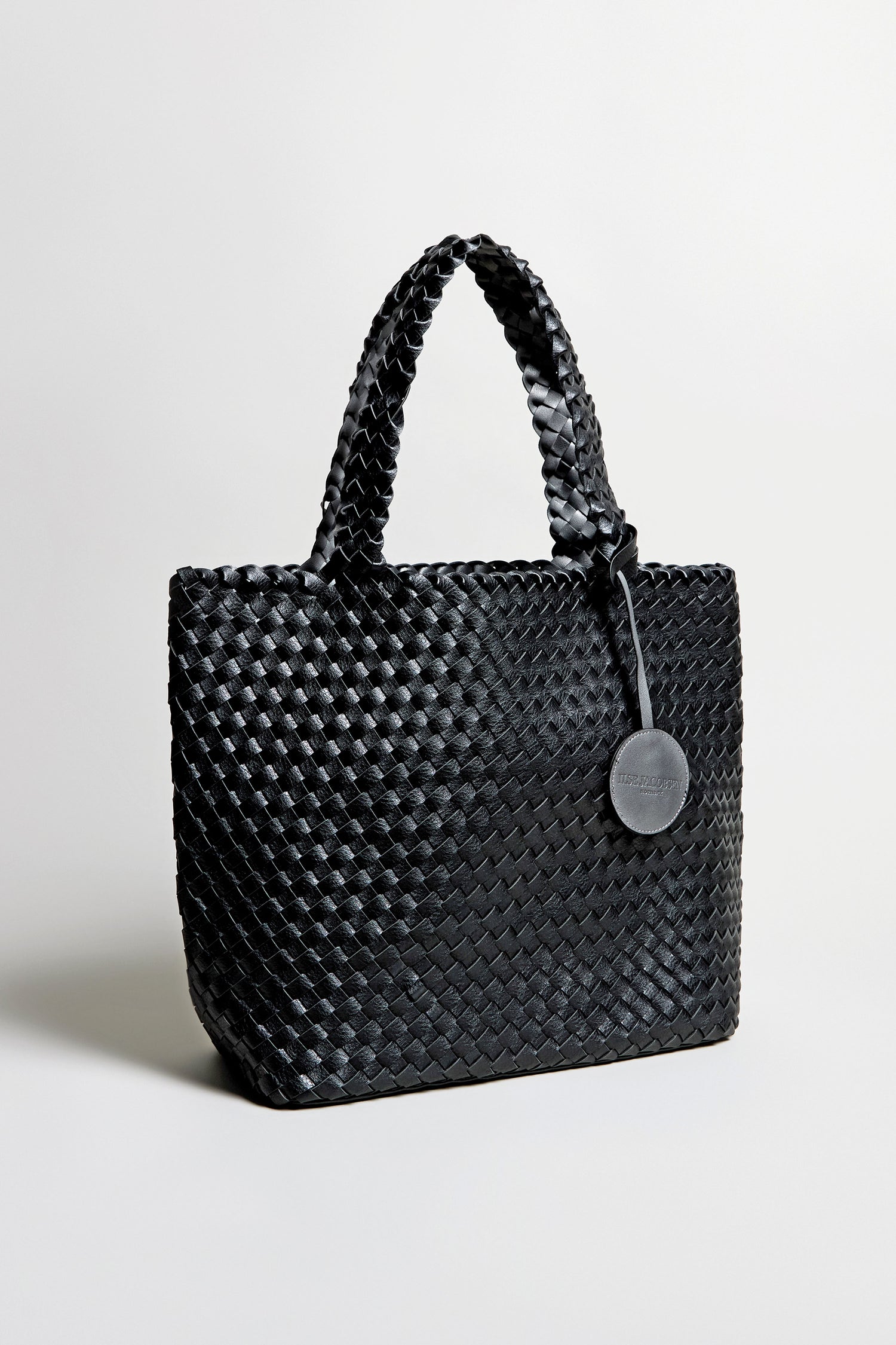 The Woven Reversible Bag features a black basket weave design, two handles, and a round leather tag, shown against a plain light background.