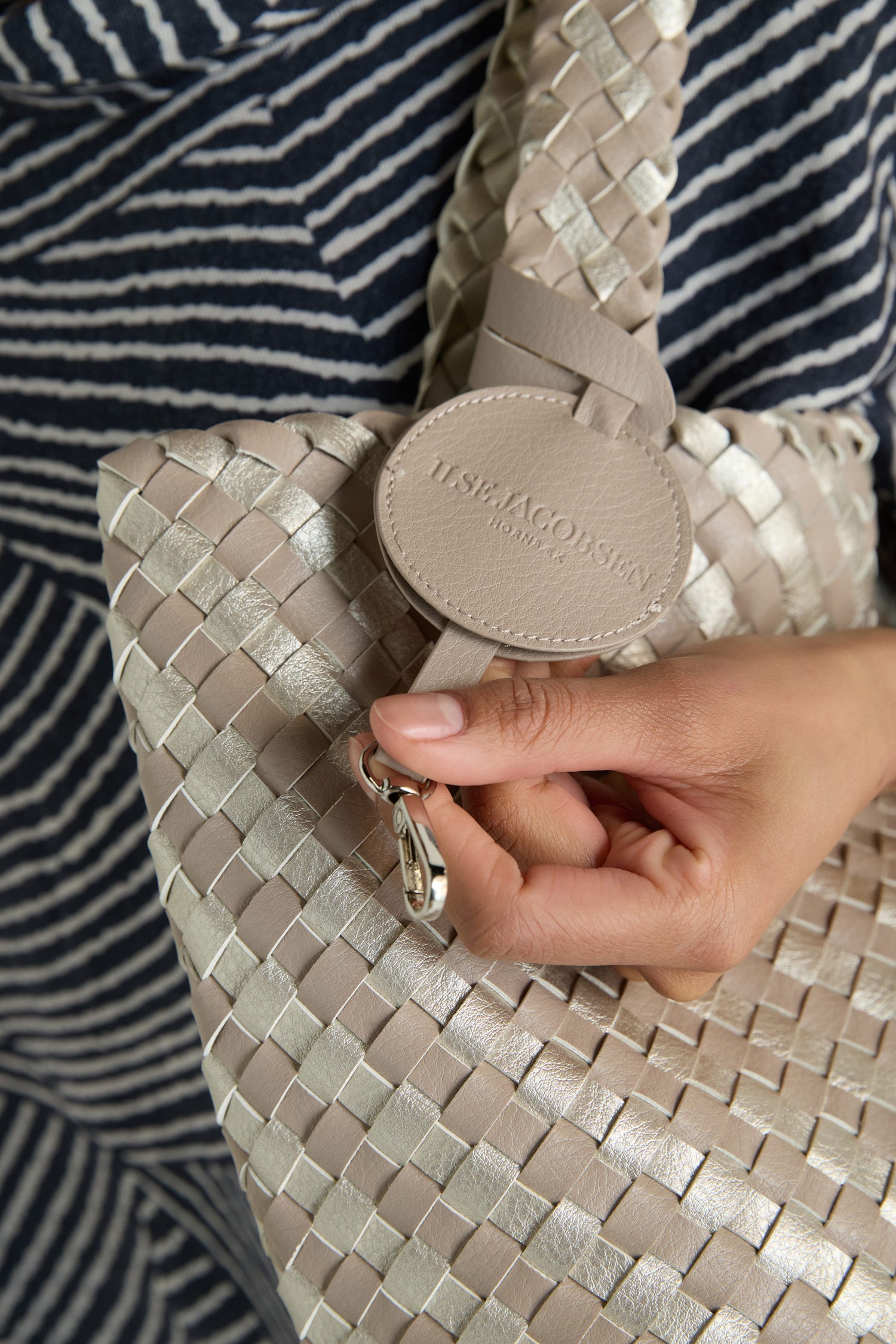 A hand holds the strap of the Woven Reversible Grey & Silver Bag, featuring a round leather "ILSE JACOBSEN Hornbæk" tag and a metal clasp near the tag.