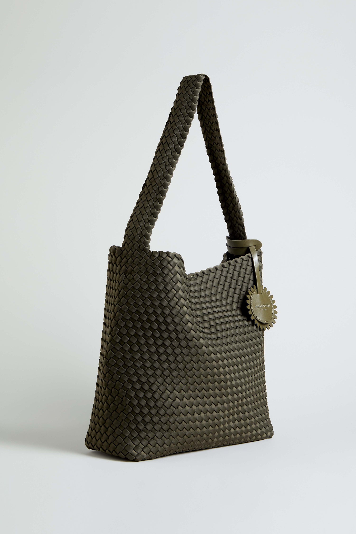 An Olive Braided Shoulder Bag with a wide strap stands upright on a plain light background, featuring a round tag hanging from the strap.