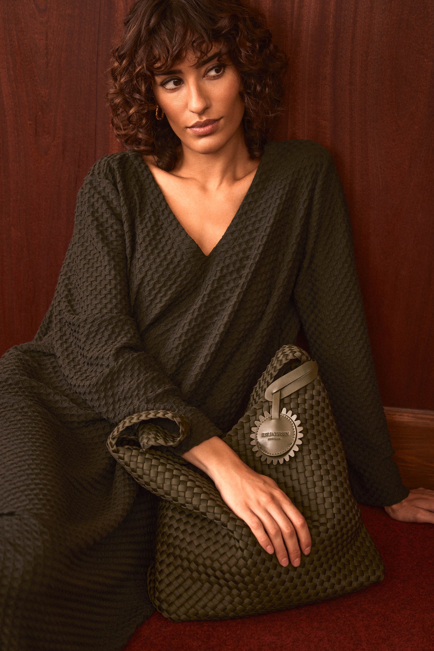 A woman with curly hair wears a textured dark green dress as she sits against a wooden wall, holding the Olive Braided Shoulder Bag—a woven green accessory accented with a metallic tag.
