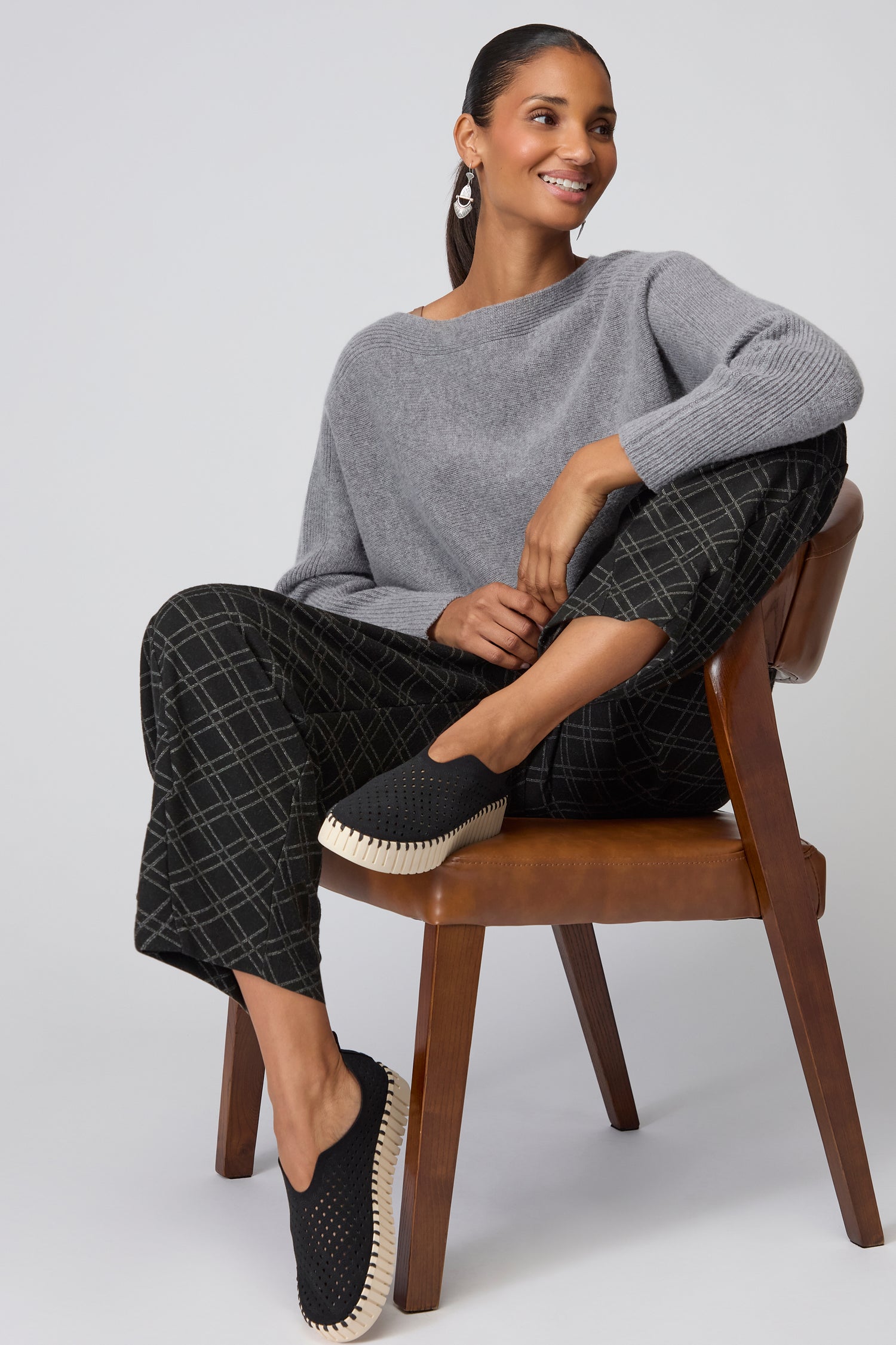 A woman sits on a wooden chair wearing a gray sweater, black checkered pants, and Black Tulip Platform Slip On Shoes. She is smiling and looking to the side.