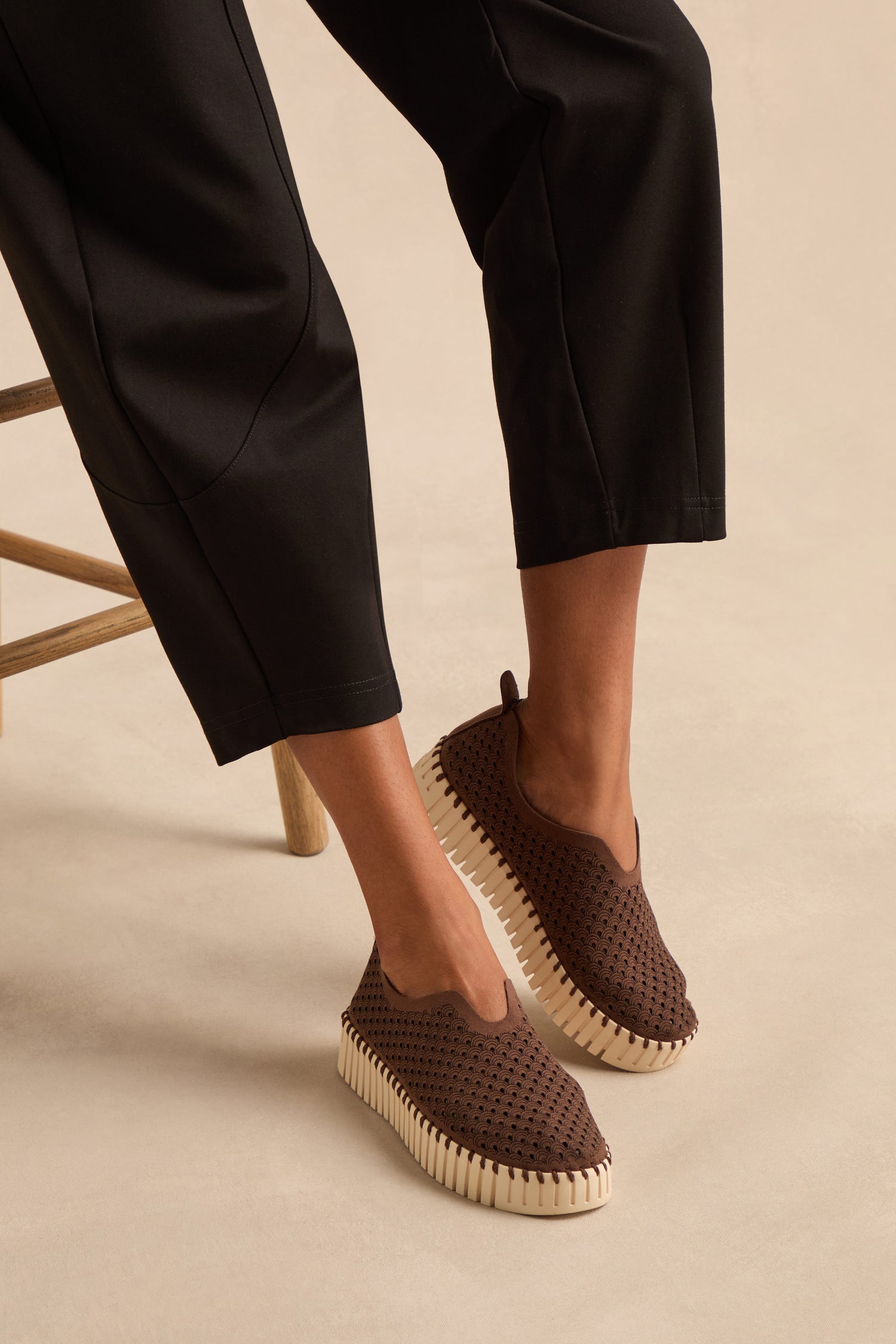 A person is seated wearing black pants and Tulip Platform Slip On Shoes—breathable slip-ons featuring brown perforated recycled microfiber uppers and thick, ridged white soles.