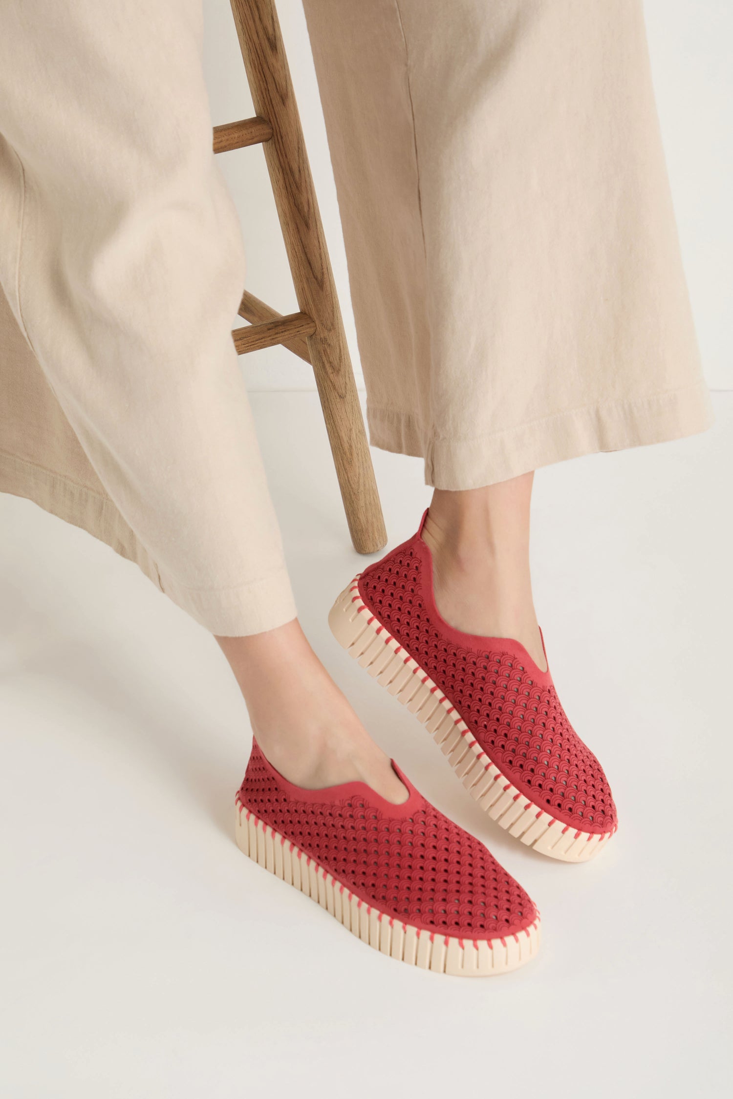A person wearing beige pants and Tulip Platform Slip on Shoes with perforated recycled microfibre uppers and thick white soles stands near a wooden stool against a white background.