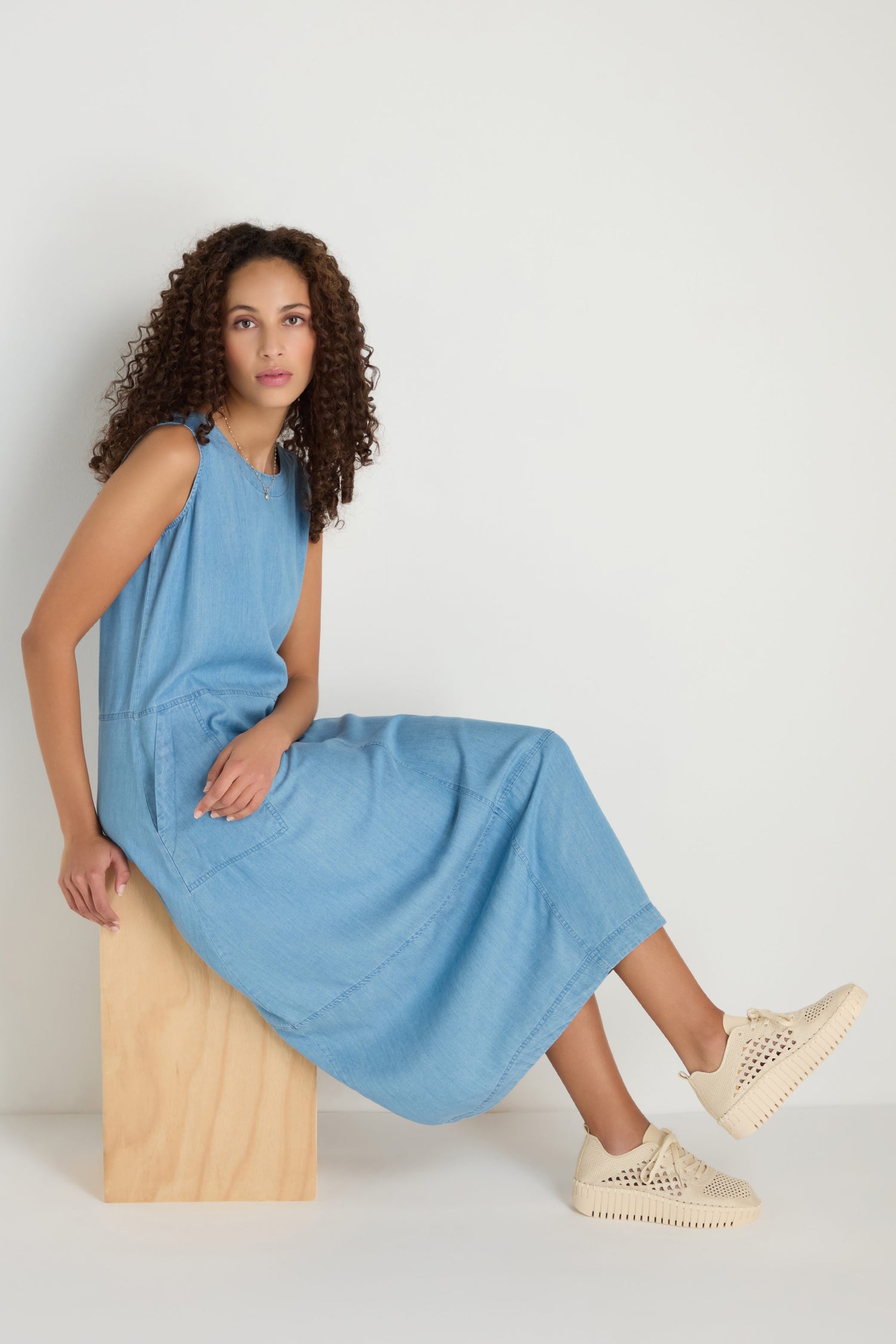 A woman with curly hair sits on a wooden block, wearing a sleeveless blue dress and Tulip Embroidered Platform Lace Up Trainers, against a plain white background.