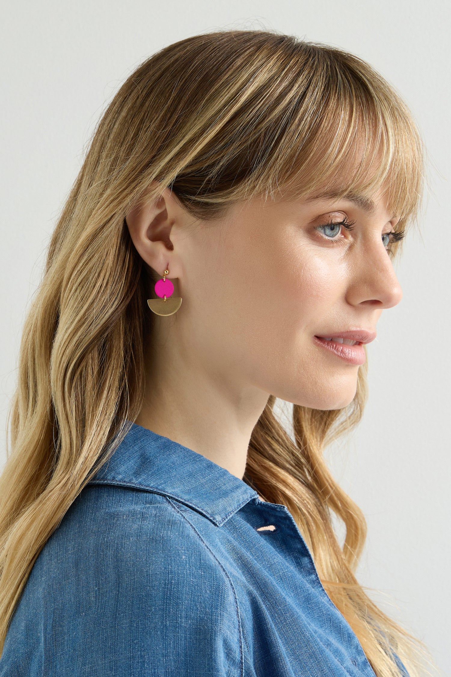 A person with long blond hair in denim wears minimalist gold and pink earrings. These elegant accessories, similar to Colour Pop Semi Circle Earrings, catch the light as they glance sideways against a plain backdrop.