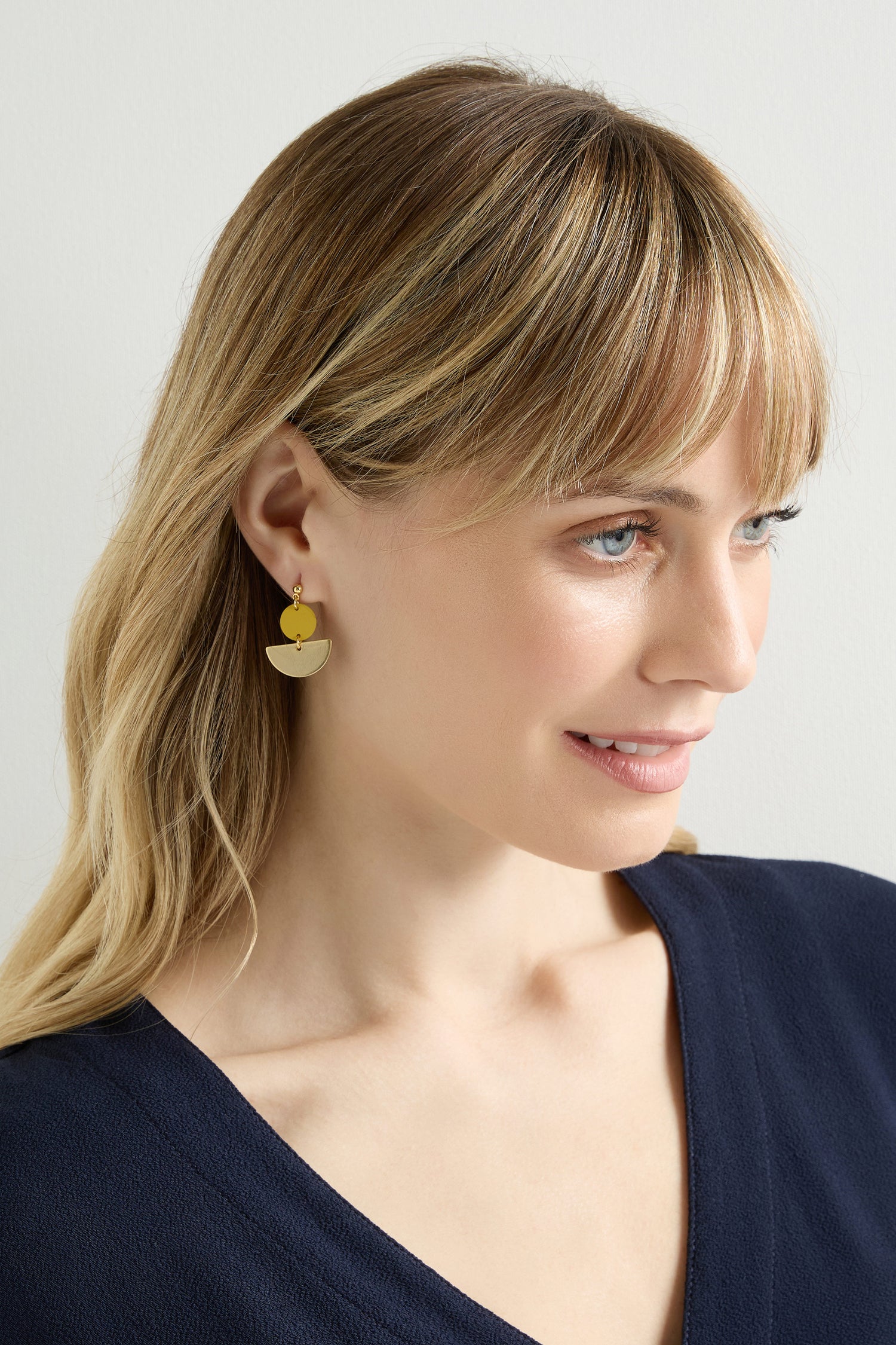 A blonde woman wears minimalist Colour Pop Semi Circle Earrings in gold and a navy V-neck top, gazing sideways against a plain background.