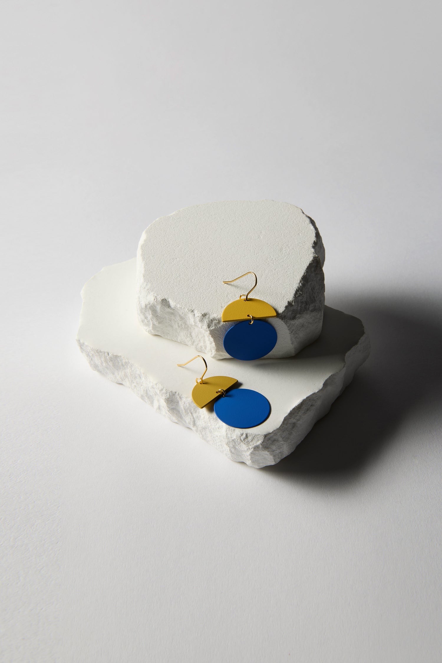 The Colour Pop Circle Drop Earrings, featuring blue circular and yellow semicircular elements, are elegantly displayed on white stone slabs against a neutral backdrop, exuding statement jewelry charm.