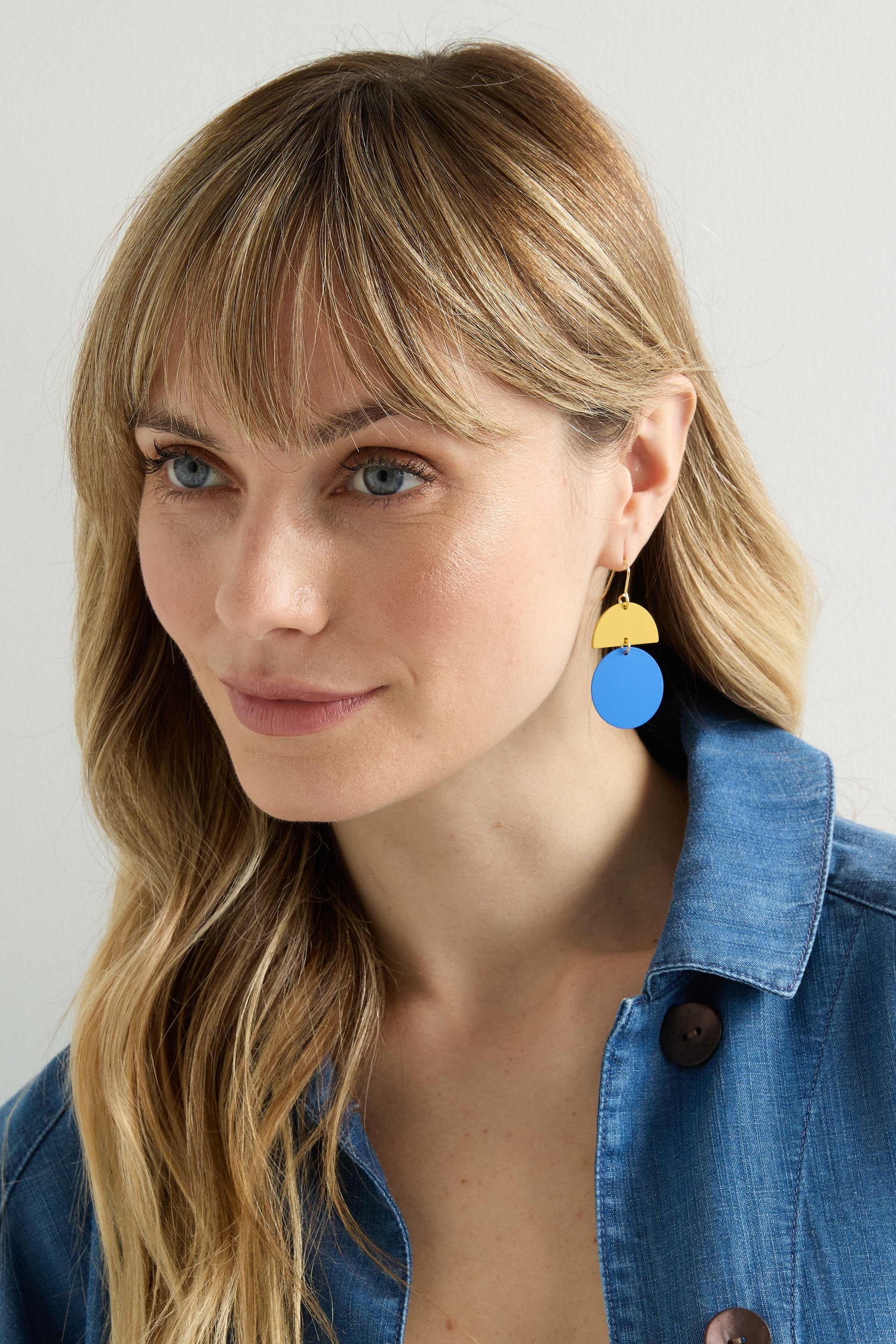 A person with long blonde hair wears a blue button-up shirt and vibrant Colour Pop Circle Drop Earrings in blue and yellow.