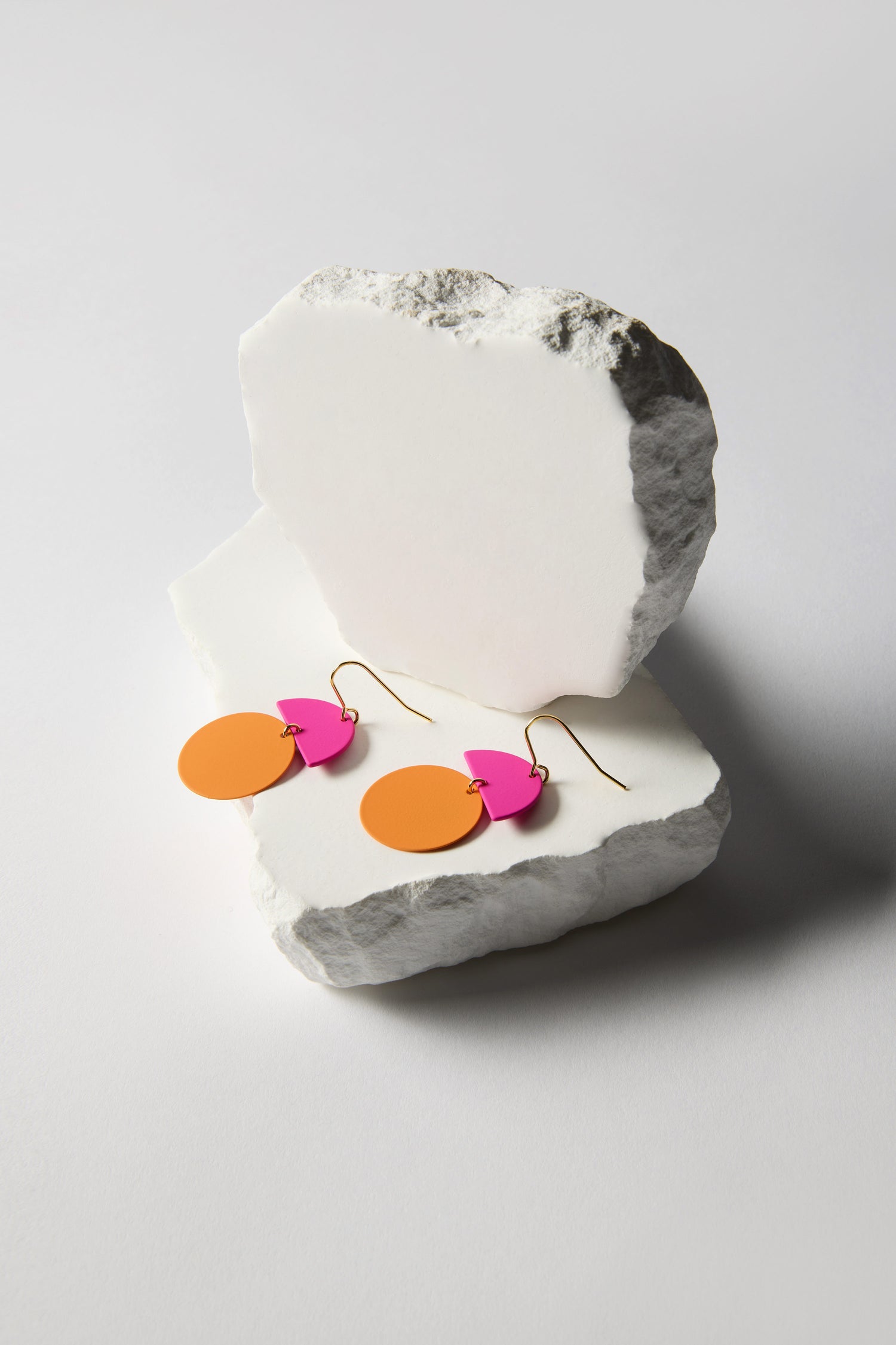The Colour Pop Circle Drop Earrings, featuring orange circles and pink abstract shapes, rest on white stone slabs against a plain background, creating a vibrant color pop that makes them standout modern pieces.