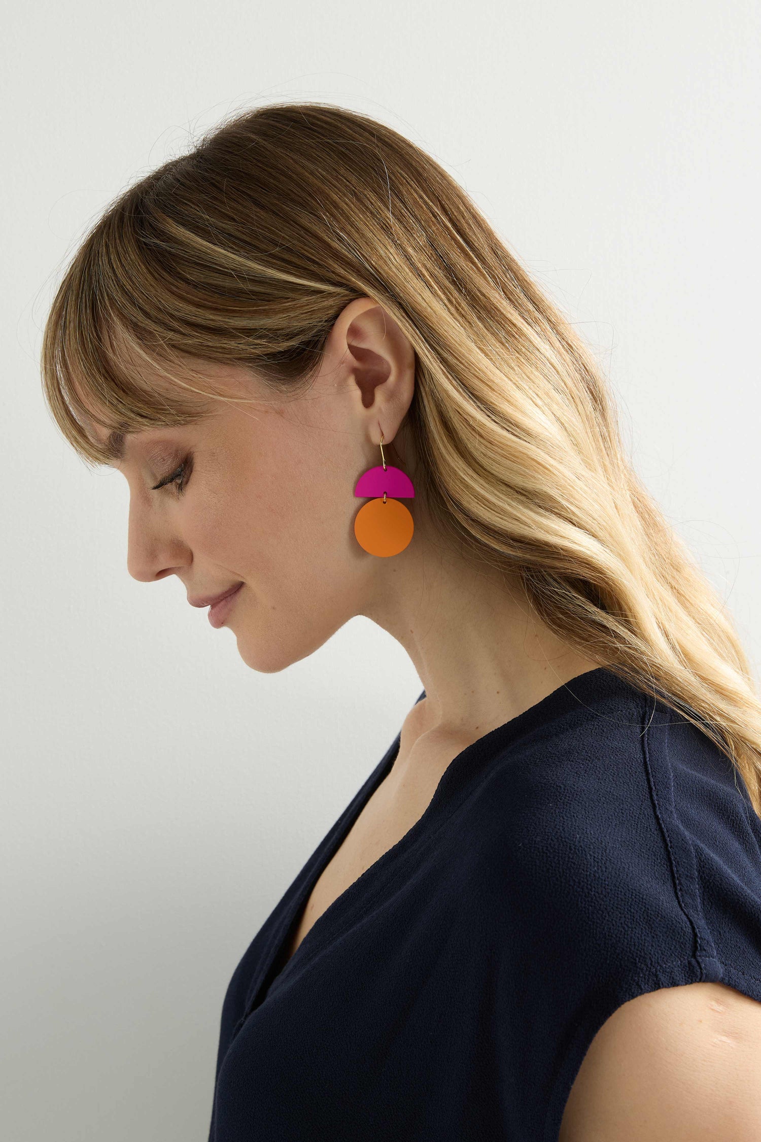 A person with wavy hair, wearing a black top and vibrant Colour Pop Circle Drop Earrings in orange and pink, is facing sideways against a plain white background.