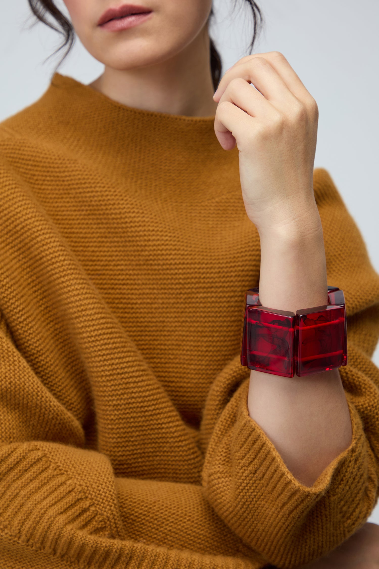 Wearing a brown knit sweater, a person displays the Ruby Resin Bracelet by Jackie Brazil on their wrist, highlighting the beauty of sustainable resin jewellery as their hand rests near their face.