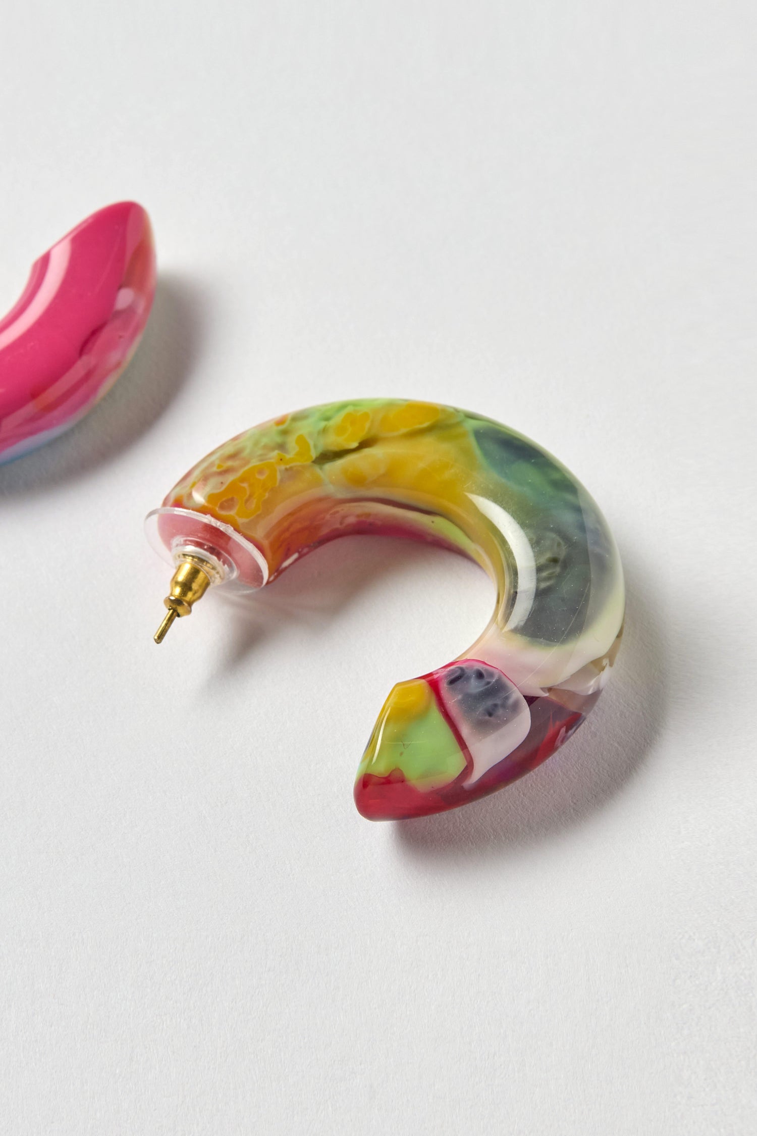 A vibrant, semi-translucent crescent-shaped resin hoop earring with a gold post rests on a light surface, showcasing the artistic style of the Kandinsky Hoop Earrings.