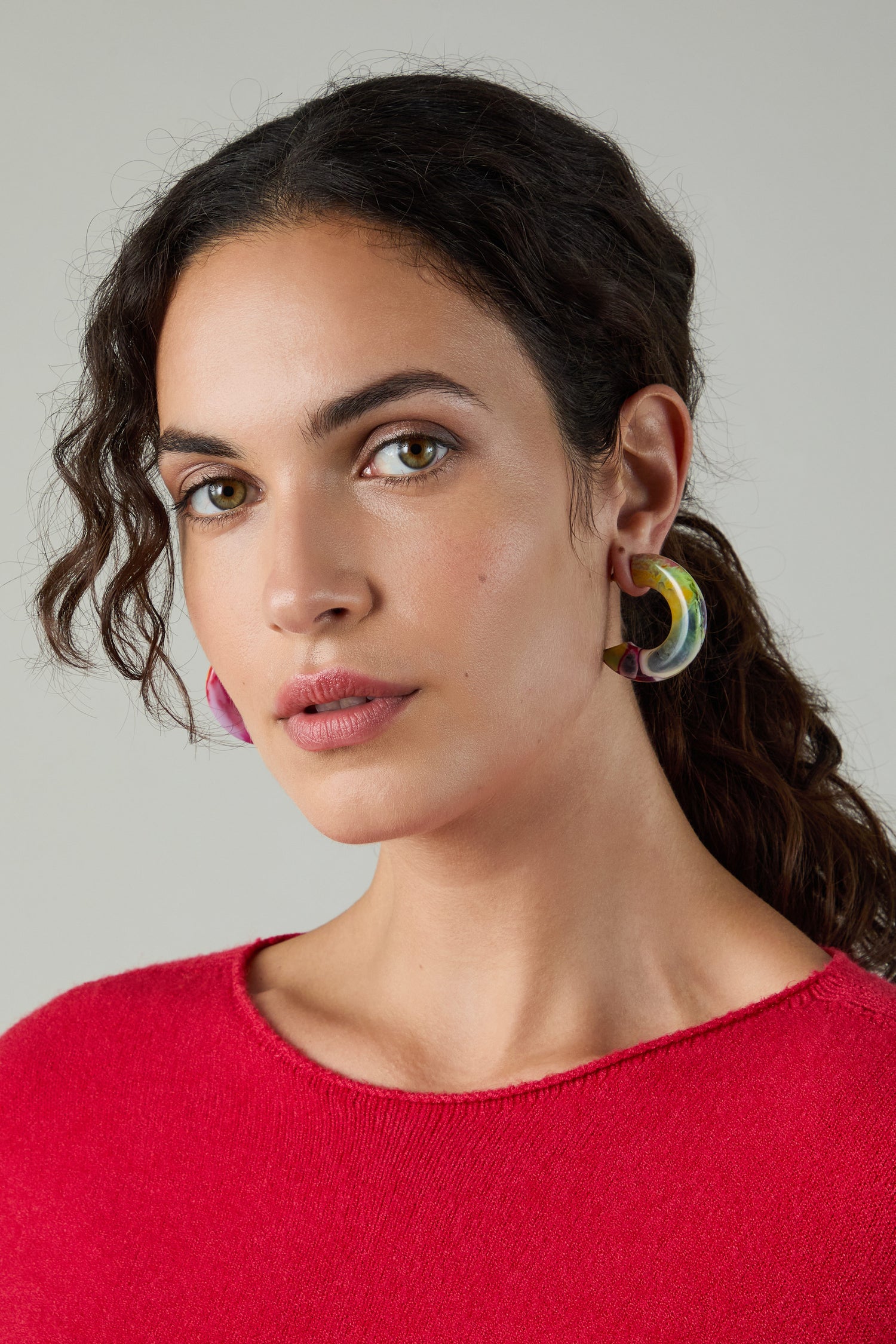 Woman with wavy dark hair wearing a red top and Kandinsky Hoop Earrings, looking at the camera against a plain light background.