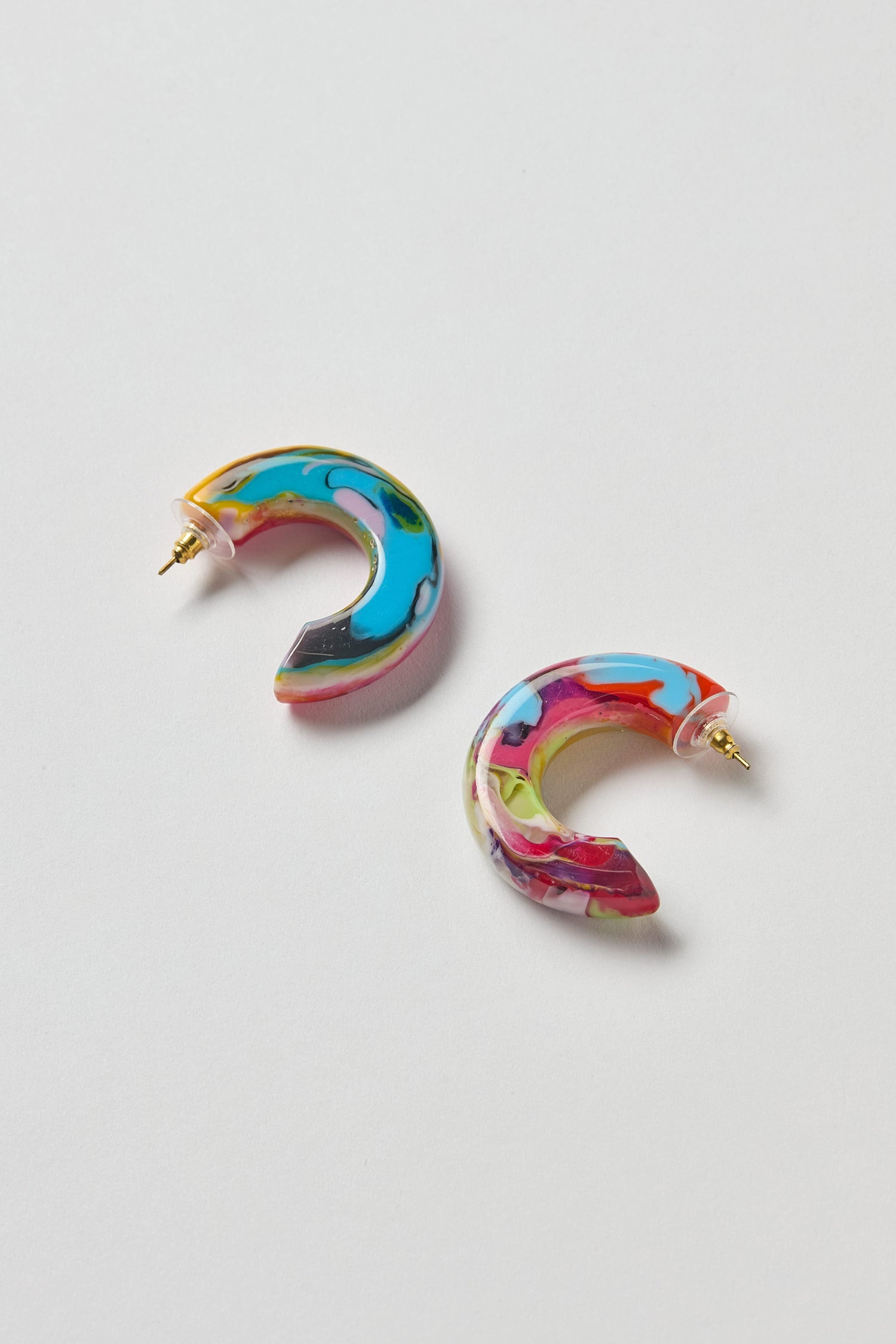 The Kandinsky Hoop Earrings feature two crescent-shaped resin hoops with a marbled blue, pink, yellow, and green pattern, set on a white background.