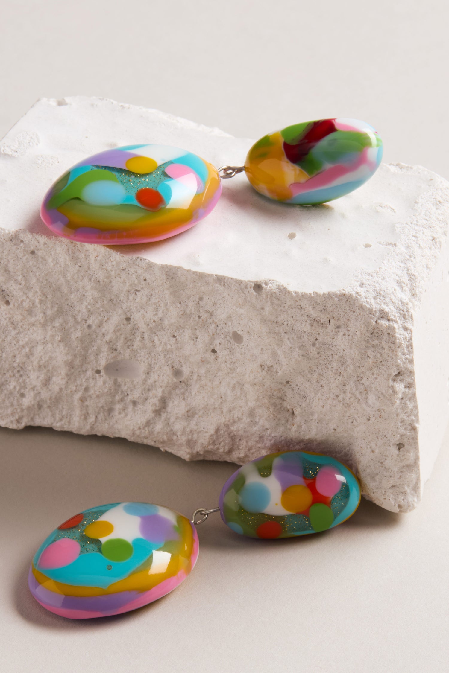 The Kandinsky Duo Pebble Earrings, featuring colorful oval shapes with abstract multicolored patterns, are displayed on a textured white stone surface.