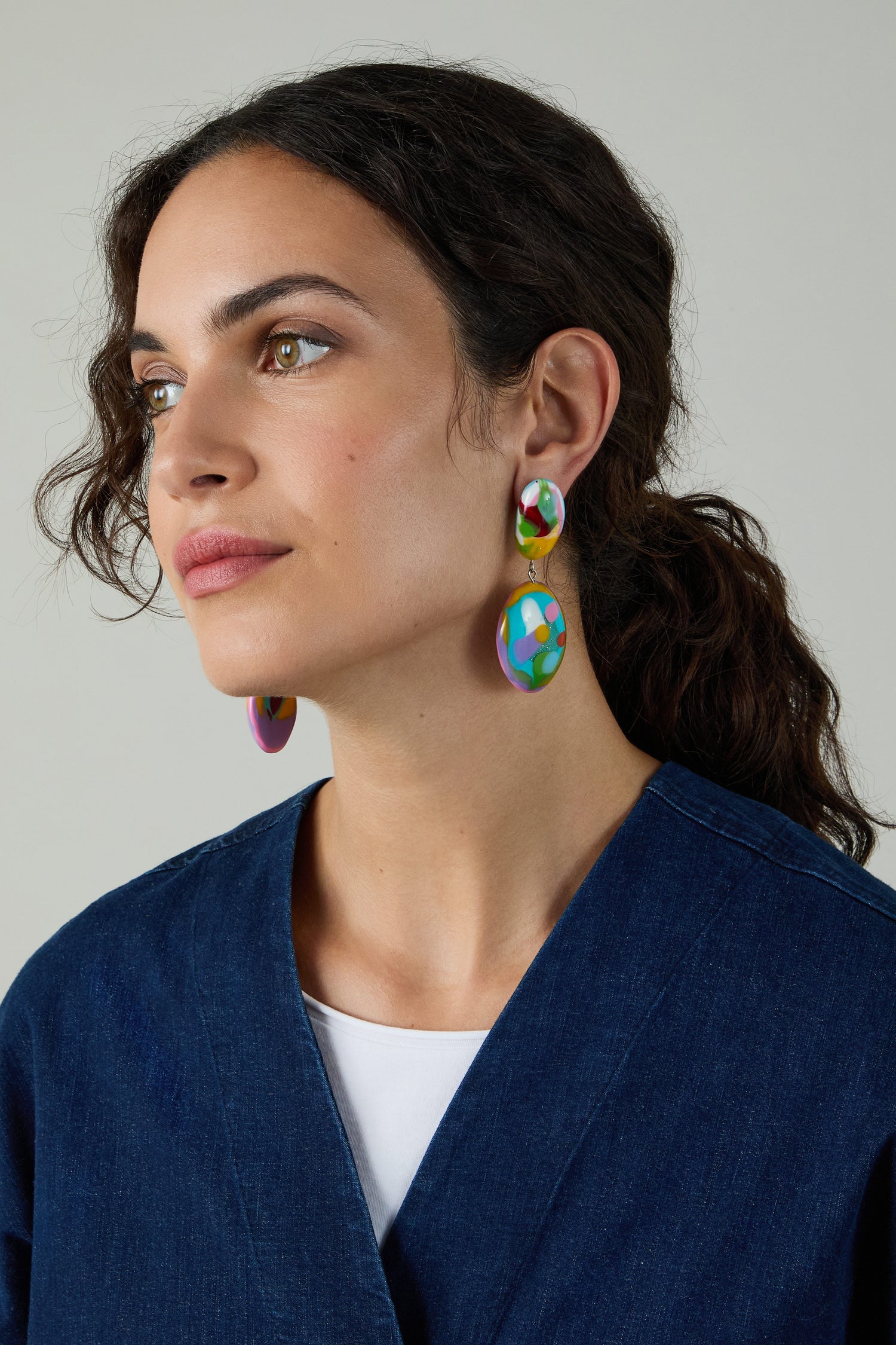 A woman with wavy dark hair in a low ponytail, wearing a blue denim top and Kandinsky Duo Pebble Earrings, looks to the side against a plain background.