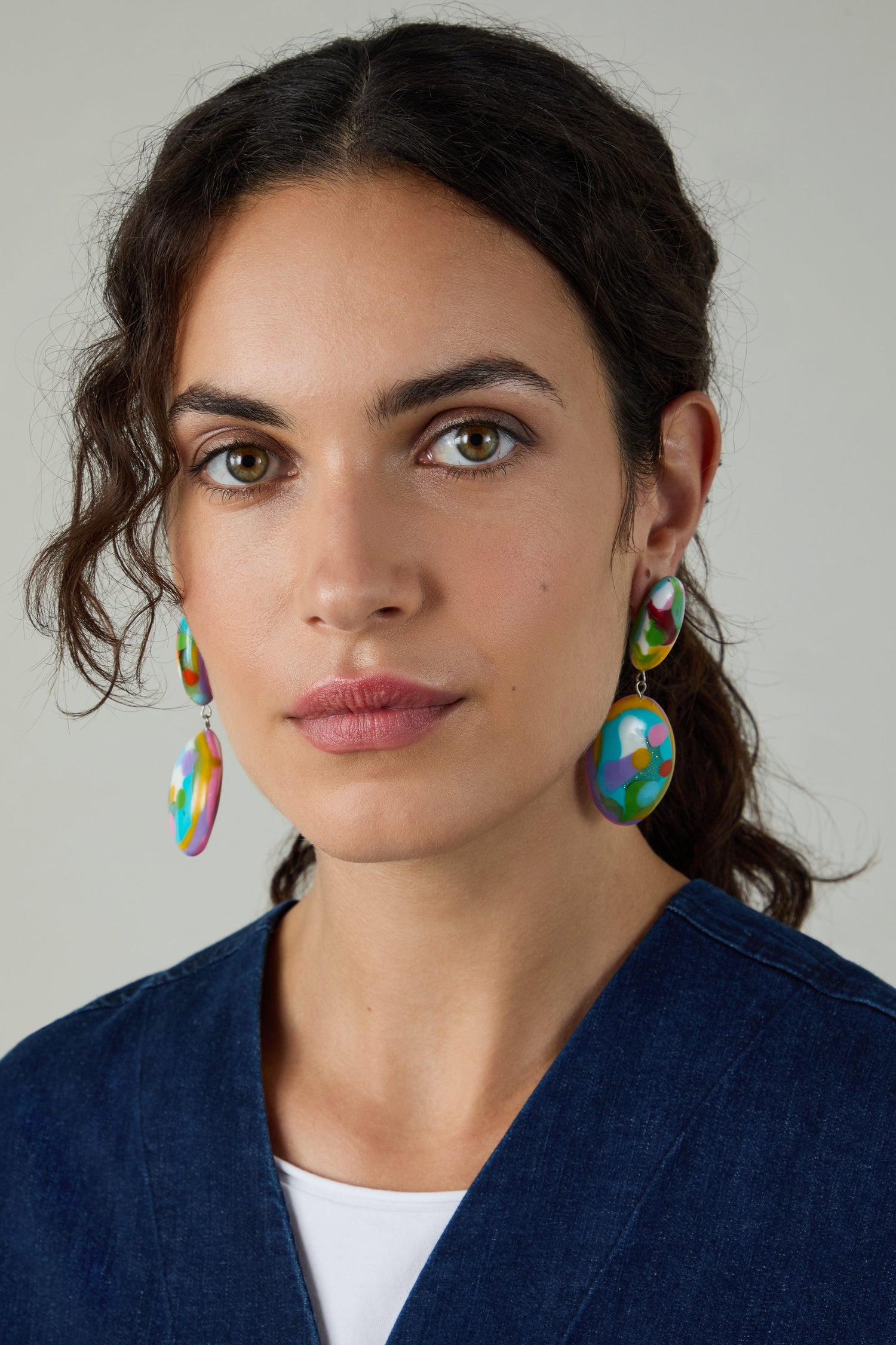 A woman with wavy brown hair wears Kandinsky Duo Pebble Earrings, a dark blue top, and a white shirt, looking directly at the camera against a plain background.