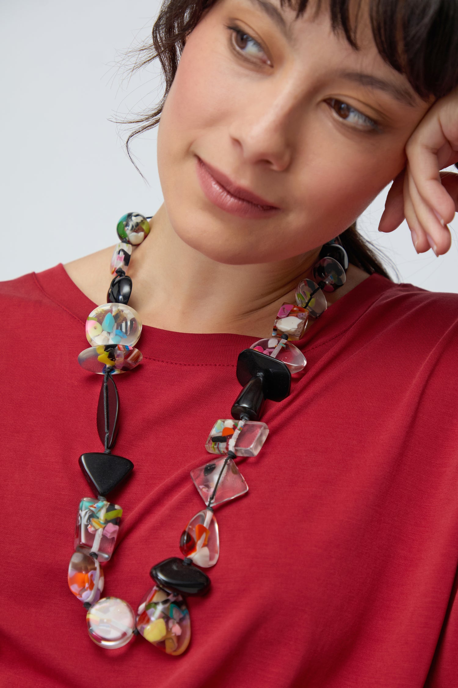 A woman in a red top rests her head on her hand, wearing the Short Multi Shapes Resin Necklace and looking slightly to the side.