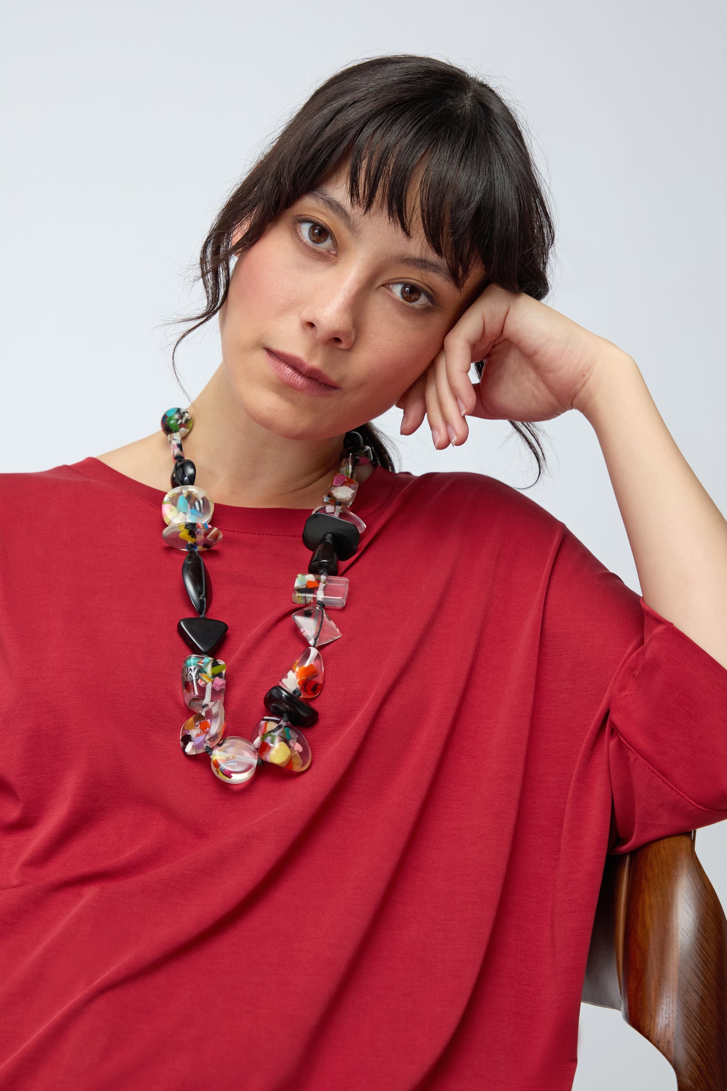 A woman with dark hair and bangs, wearing a loose red top and the Short Multi Shapes Resin Necklace, sits with her head resting on her hand, looking at the camera.