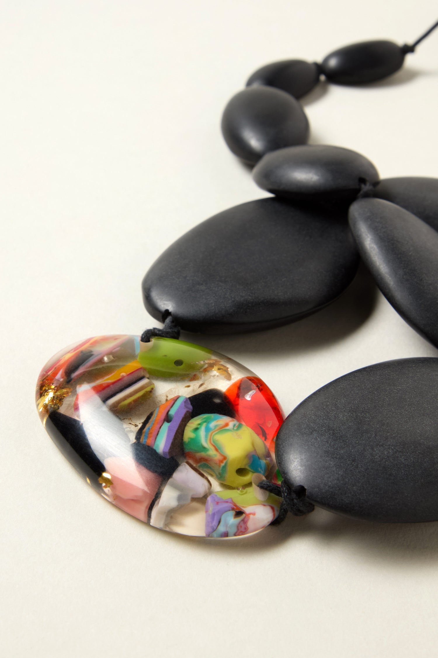The Riverstones Resin Pebble Necklace by Jackie Brazil features large matte black oval beads and a unique multicolored abstract pendant, making it a bold statement of sustainable jewellery.