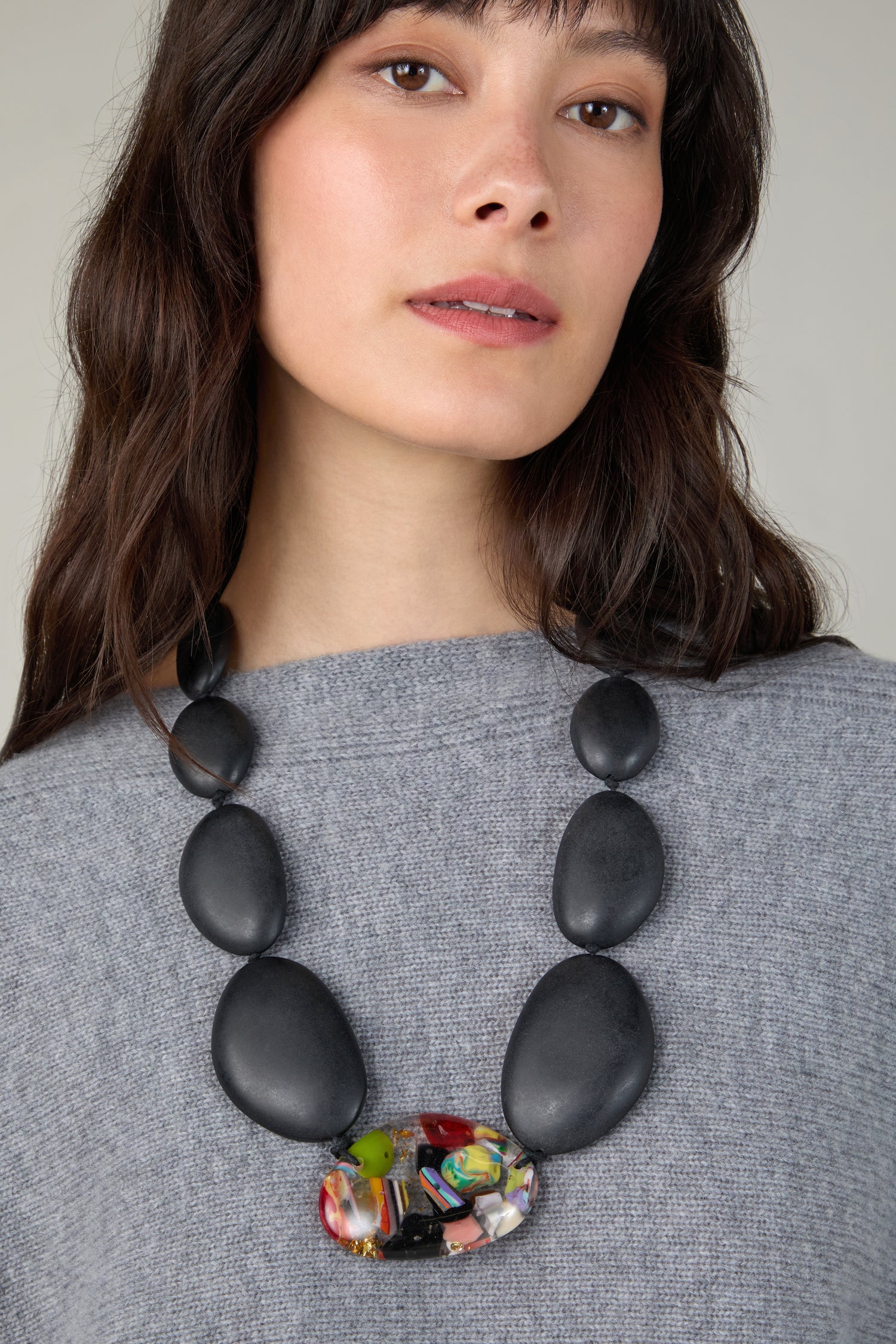 A woman with long dark hair wears a grey top and the Riverstones Resin Pebble Necklace featuring large black beads and a multicolored central bead, highlighting her love for sustainable jewellery.
