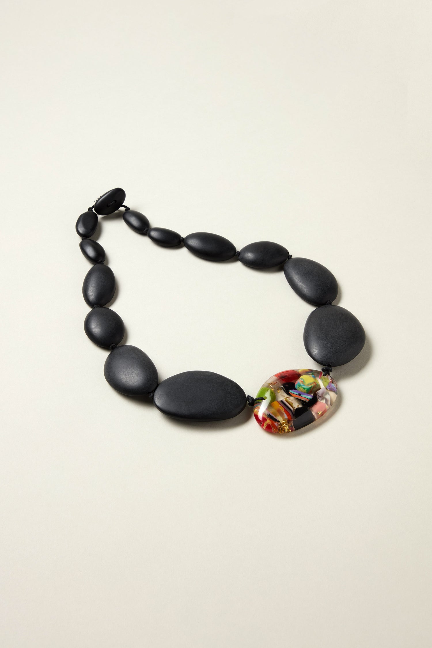 The Riverstones Resin Pebble Necklace features large matte black oval beads and a striking multicolored centerpiece, displayed on a plain light background.