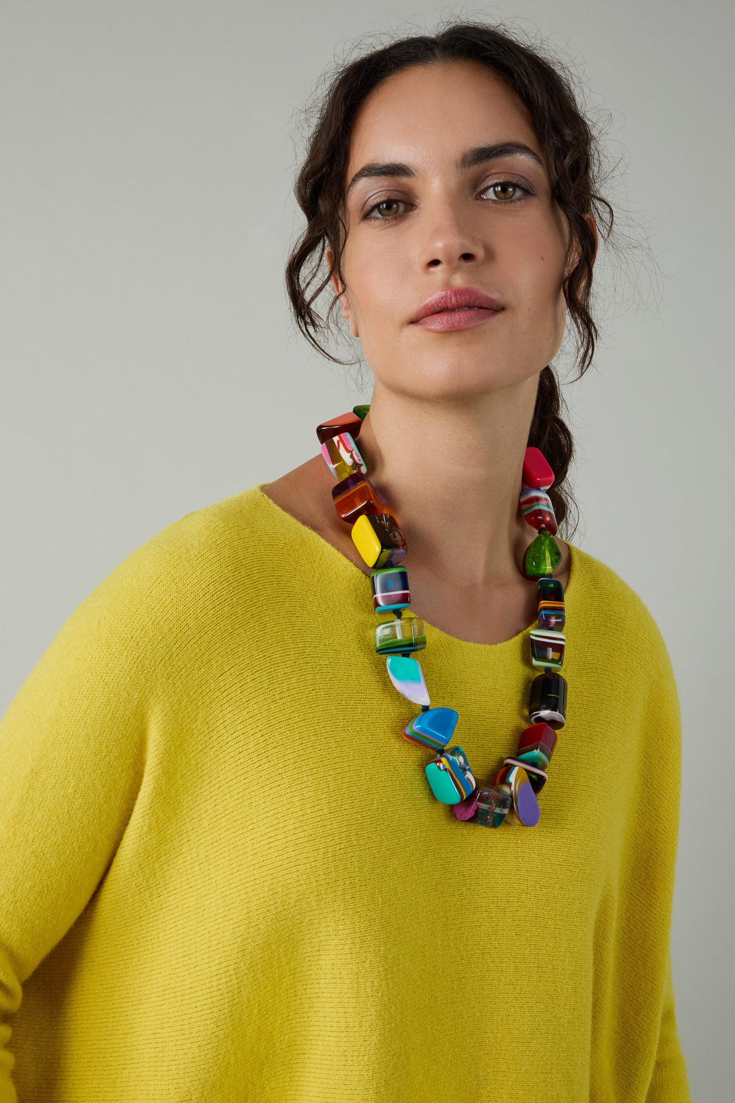 A woman in a yellow sweater wears the Kandinsky Pop Art Pebble Necklace, a chunky, handcrafted resin piece inspired by pebble designs, standing against a plain gray background.