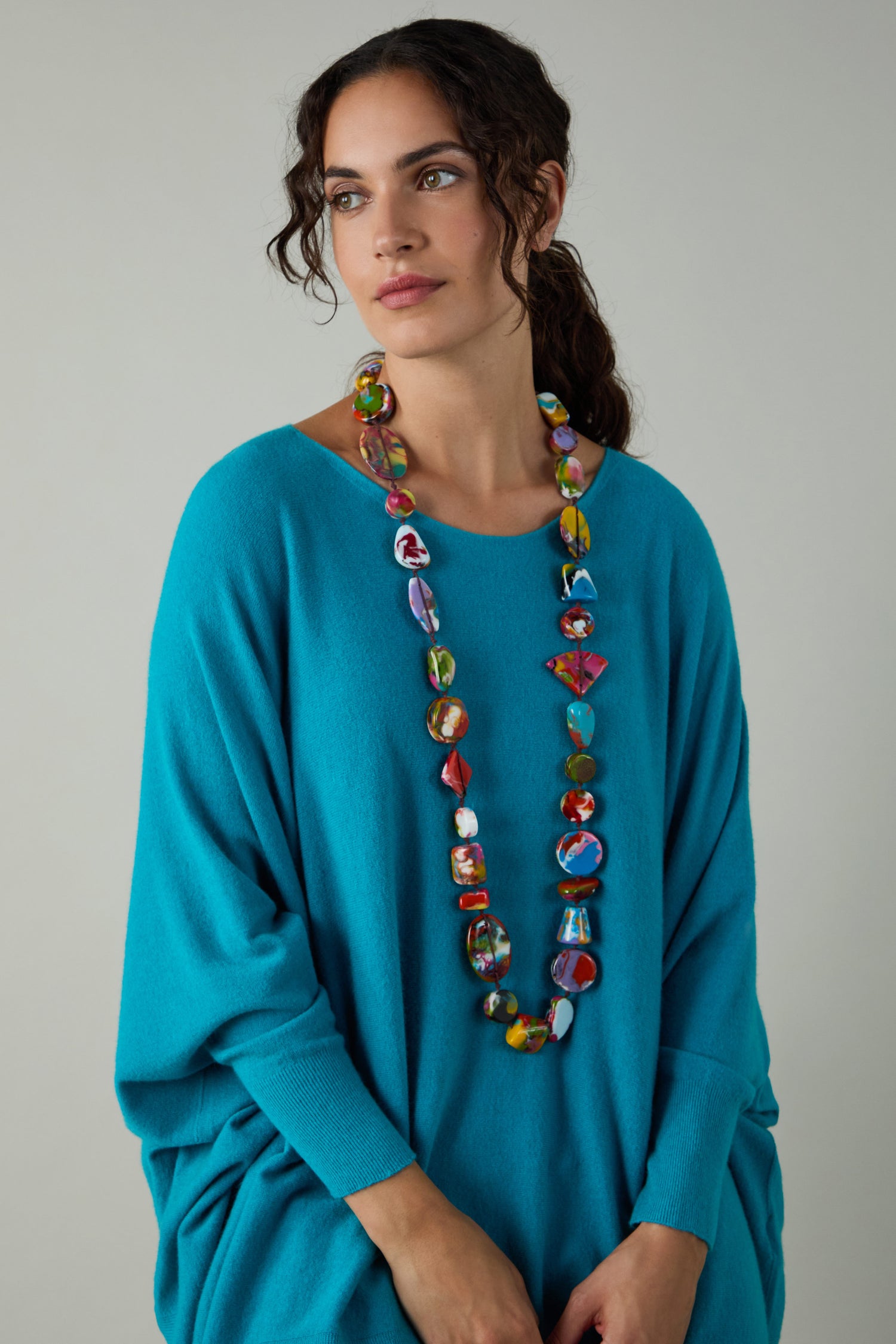 A woman in a loose teal sweater wears the Long Kandinsky Pebble Resin Necklace—a colorful, sculptural strand—while standing against a plain light gray background.