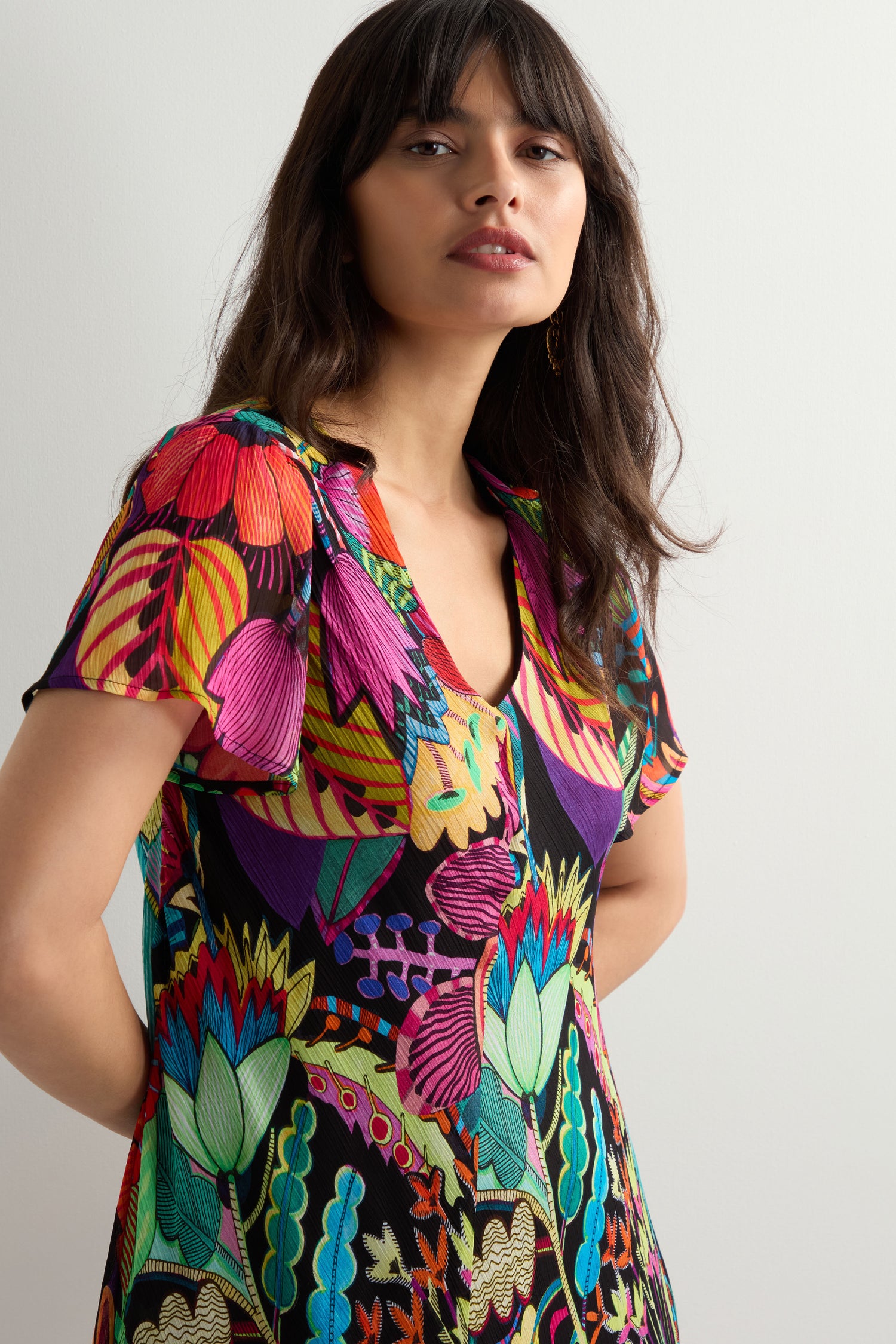 A woman with long dark hair models the Fantasy Forest Georgette Bubble Dress with tropical flora and geometric patterns in a relaxed, oversized fit, standing before a plain light background.