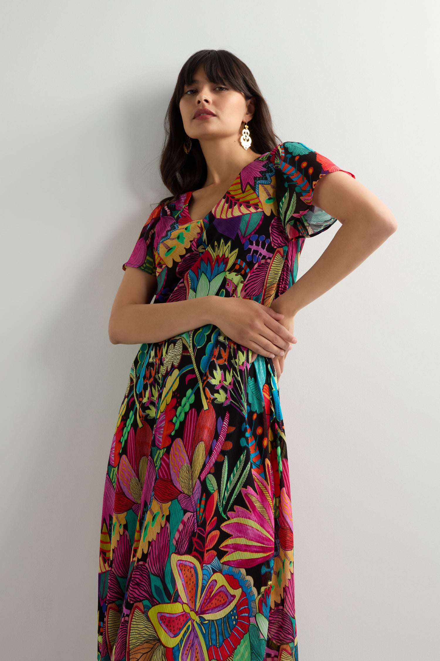 A woman in the Fantasy Forest Georgette Bubble Dress with tropical flora patterns and gold earrings stands against a white background, gazing upward in a relaxed oversized fit.
