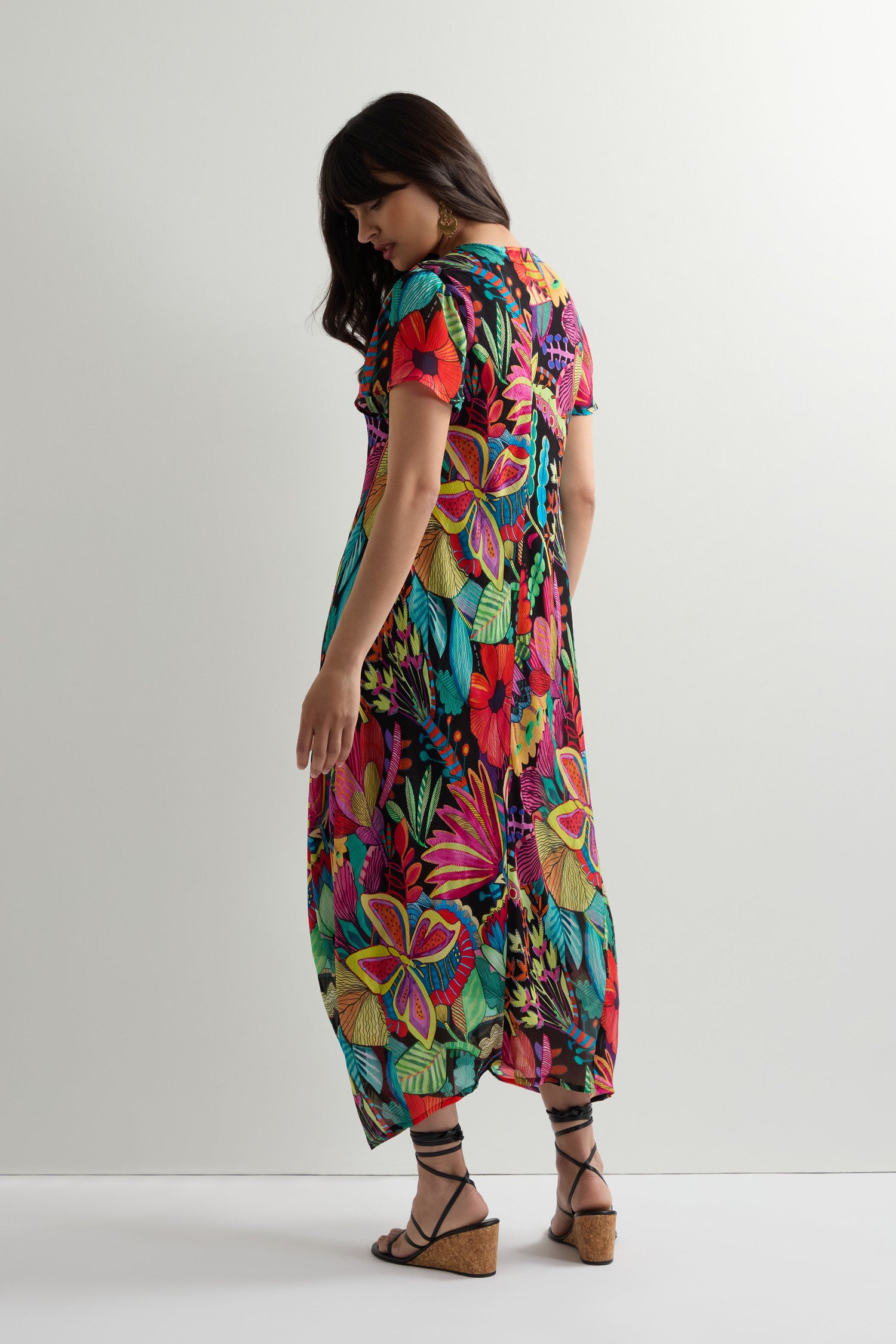 A woman poses against a plain background wearing the Fantasy Forest Georgette Bubble Dress, featuring a colorful floral print, relaxed oversized fit, and short sleeves, styled with wedge sandals.