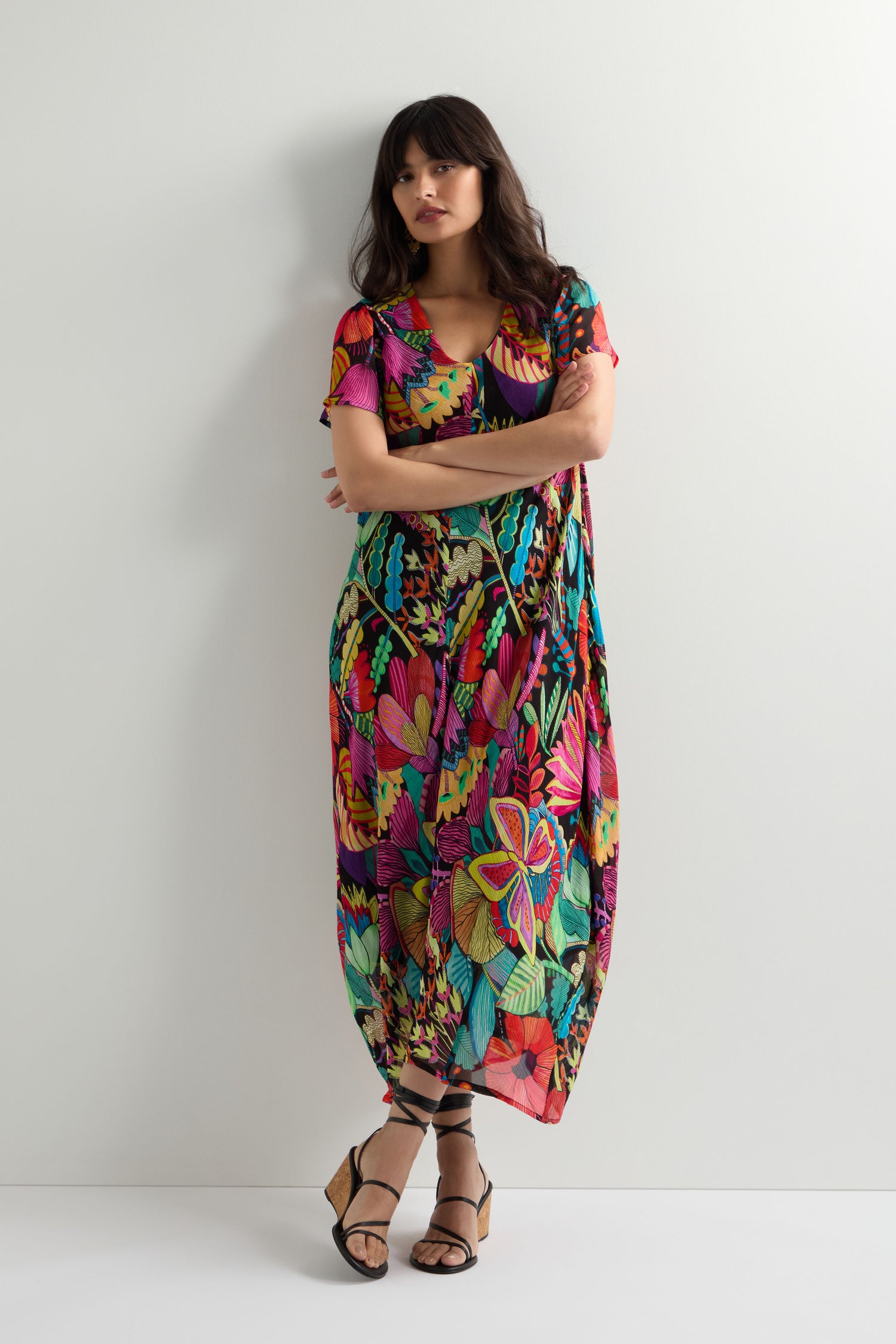 A woman stands against a white wall, arms crossed, wearing the Fantasy Forest Georgette Bubble Dress in tropical flora with a relaxed oversized fit and black strappy sandals.