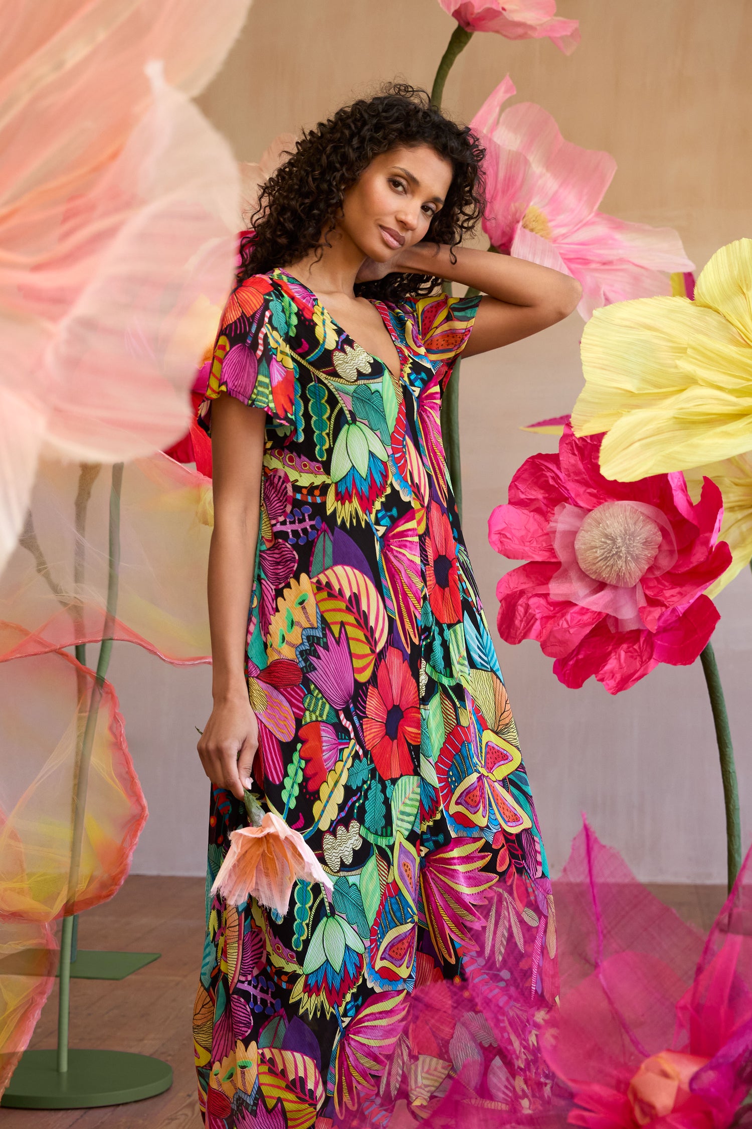 A woman wearing the Fantasy Forest Georgette Bubble Dress with a relaxed, oversized fit stands among vibrant tropical artificial flowers, holding one blossom and looking at the camera.