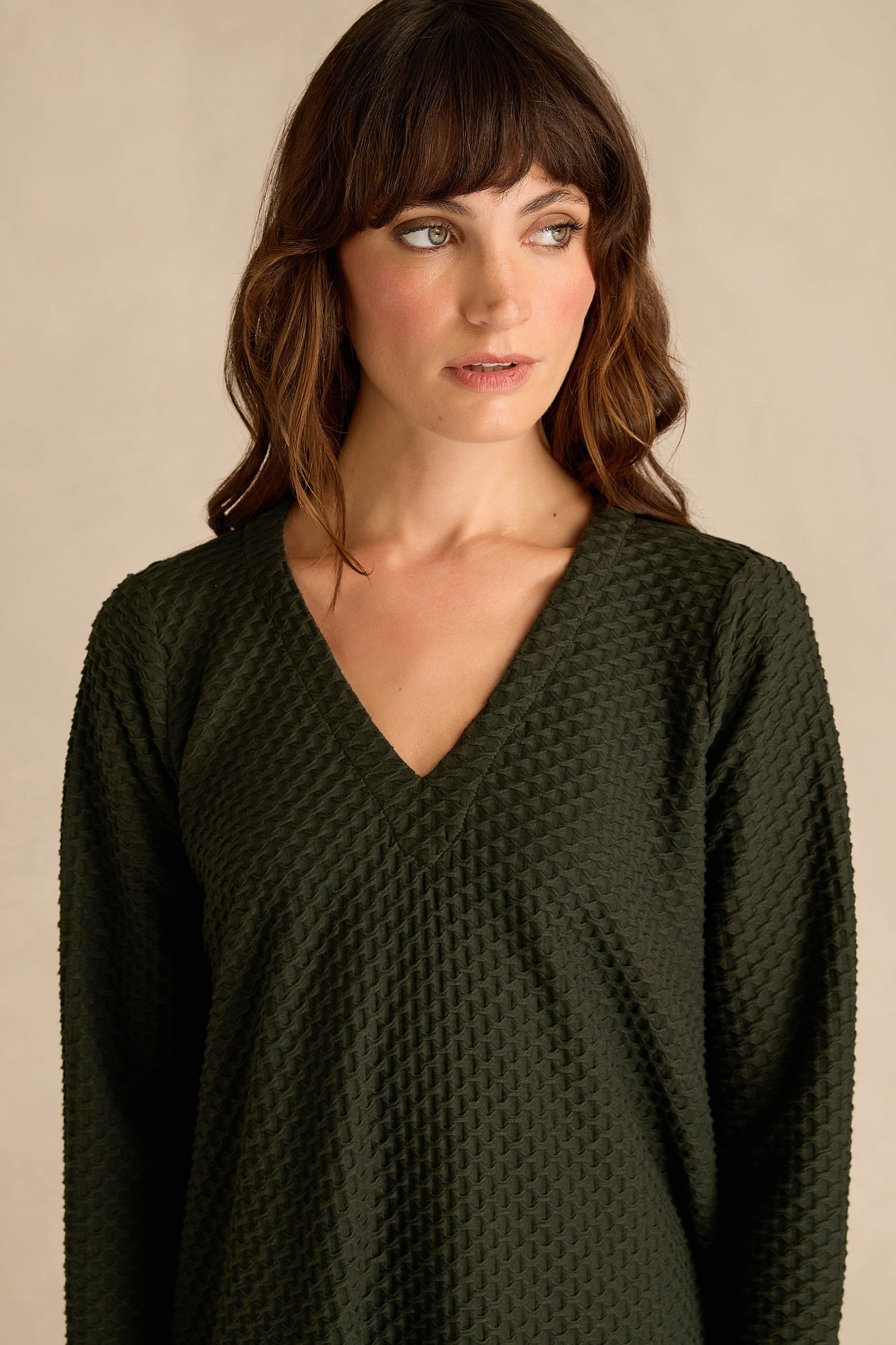 A woman with wavy brown hair is wearing the Waffle Textured Jersey Drape Dress in deep pine green, featuring a draped front and V-neck with long sleeves, as she gazes slightly to her left against a neutral background.