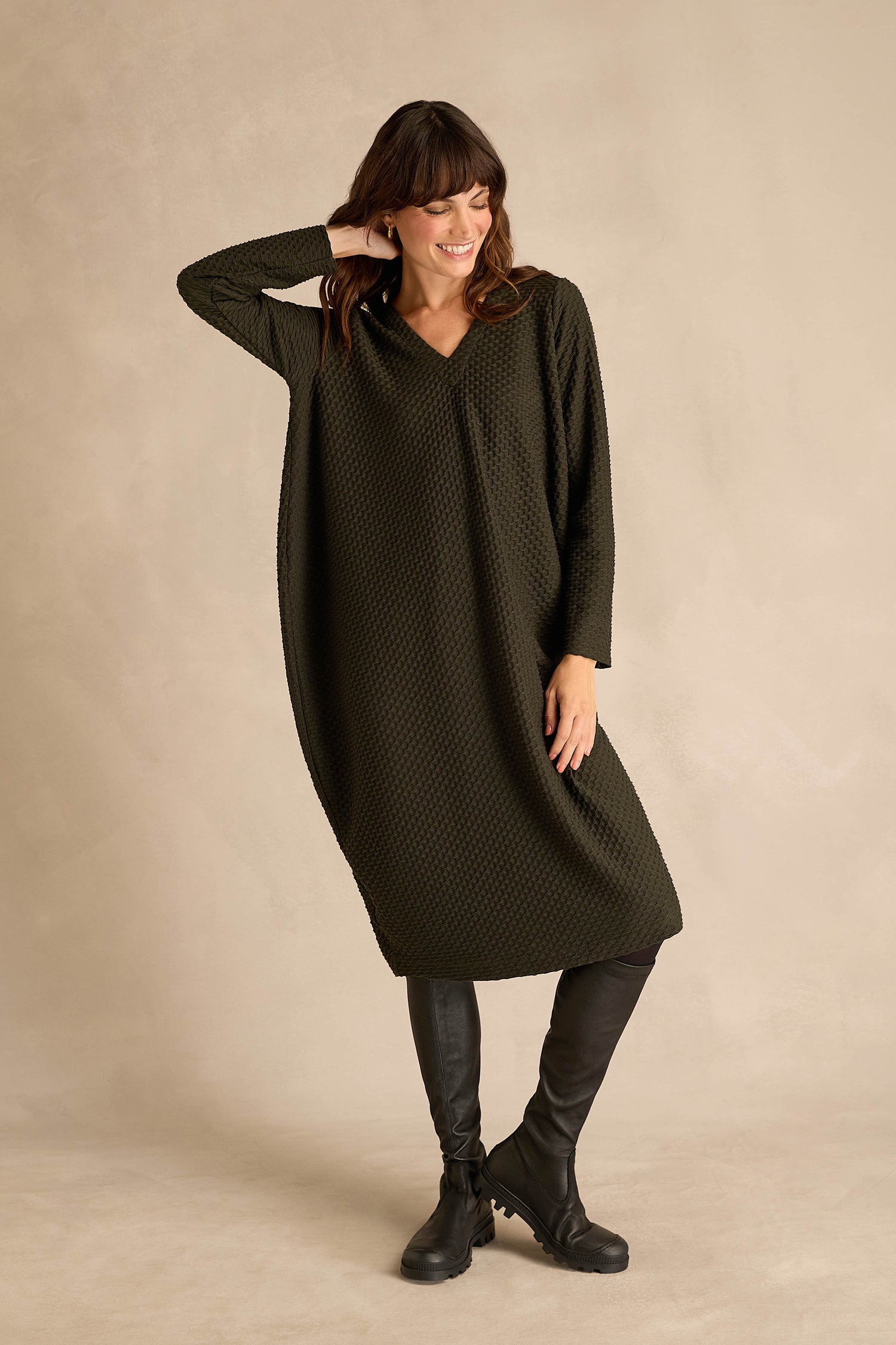 A woman stands against a light background, smiling with one hand touching her head, wearing a loose, pine green Waffle Textured Jersey Drape Dress with long sleeves and knee-high black boots.