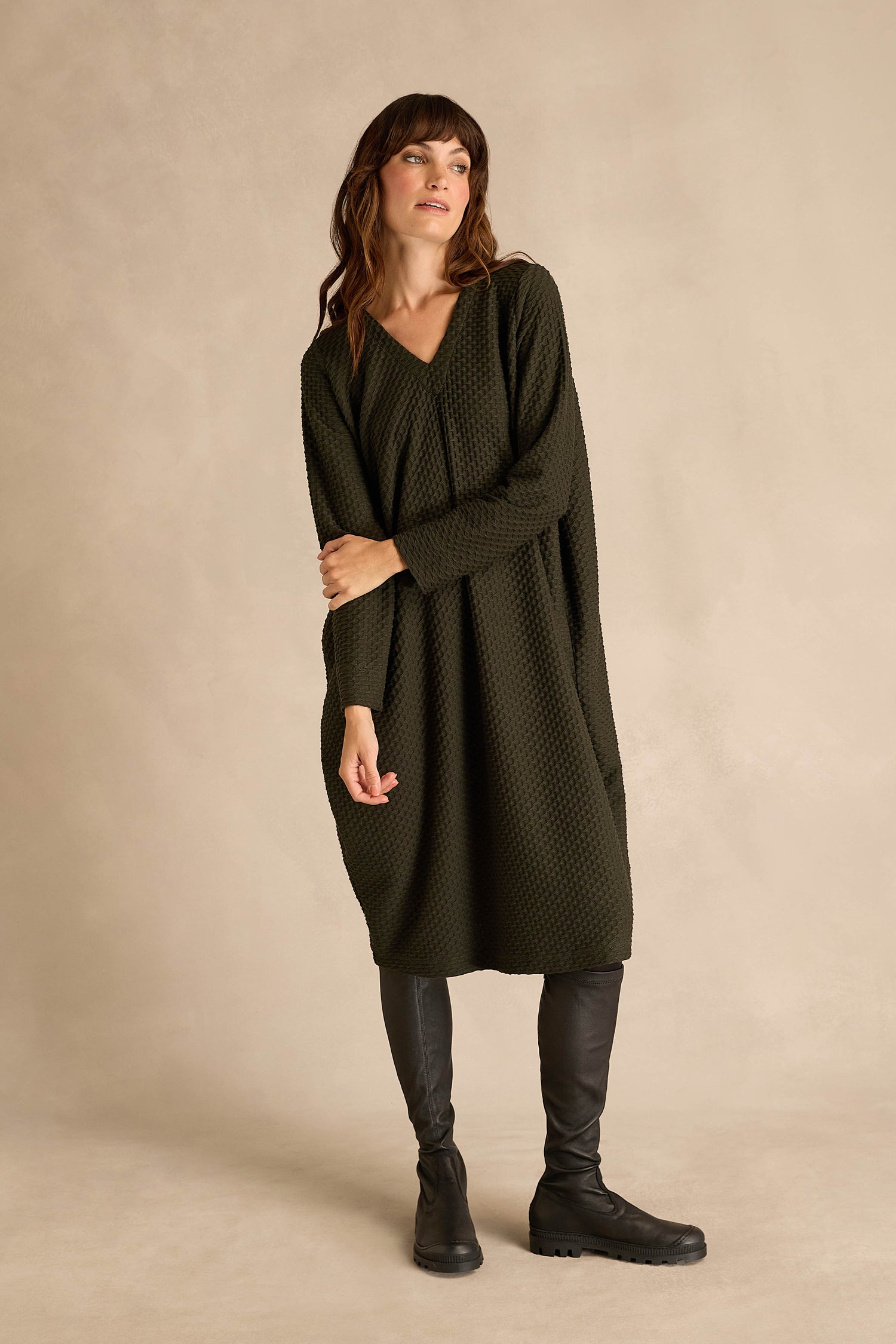 Wearing the Waffle Textured Jersey Drape Dress in deep pine green with black knee-high boots, a woman stands against a beige background.