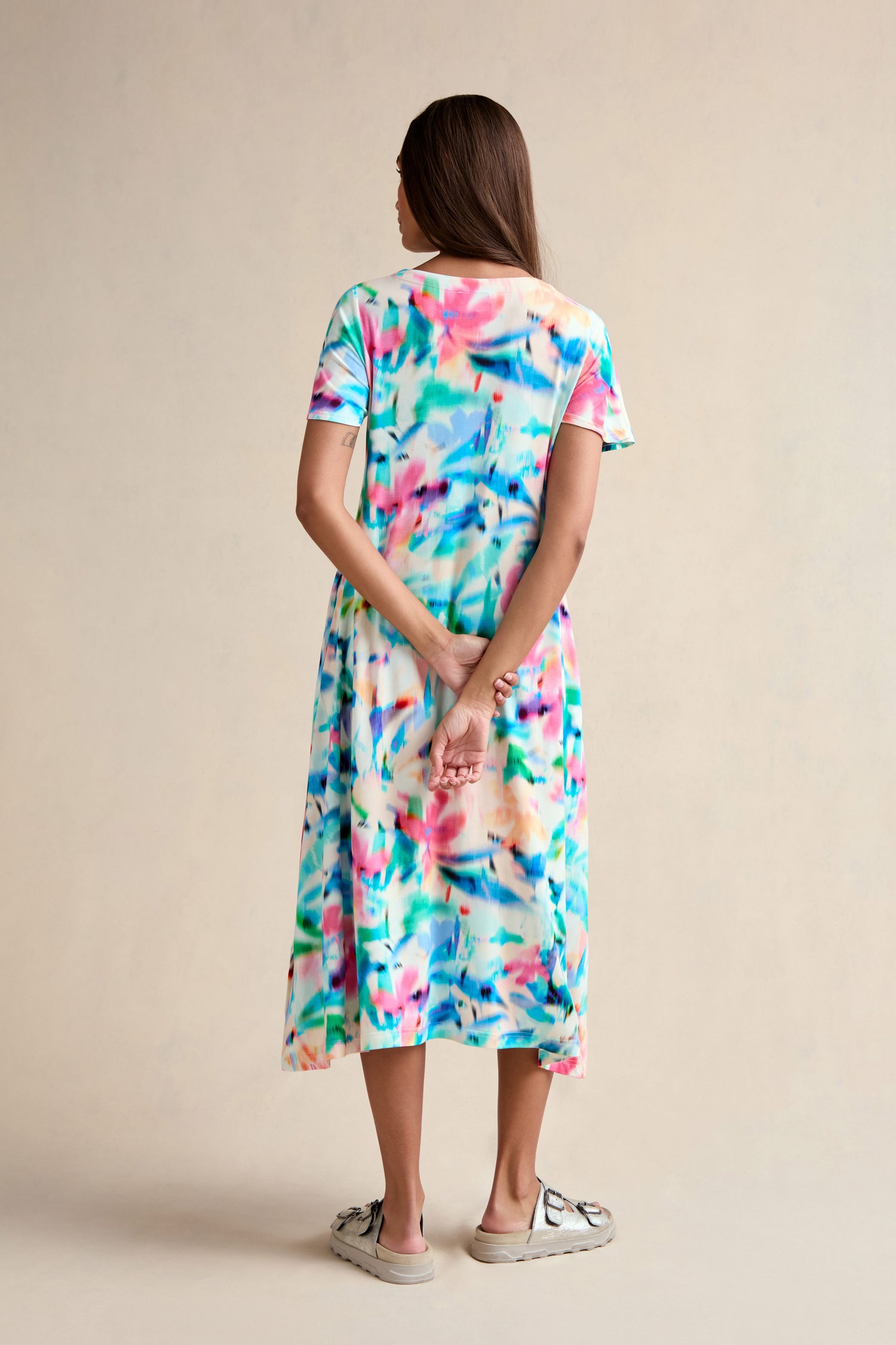 A woman with long brown hair, facing away, wears a Petal Haze Jersey Flare Dress—a colorful patterned dress—and white sneakers, her hands clasped behind her back.