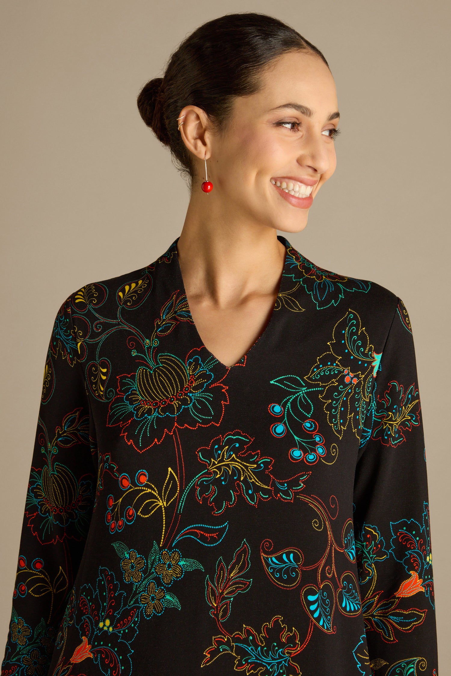 A woman smiles, glancing to the side, wearing a Bright Dotted Floral Jersey Dress featuring a V-neckline and vibrant patterns. She pairs it with red earrings, her hair styled in a bun.
