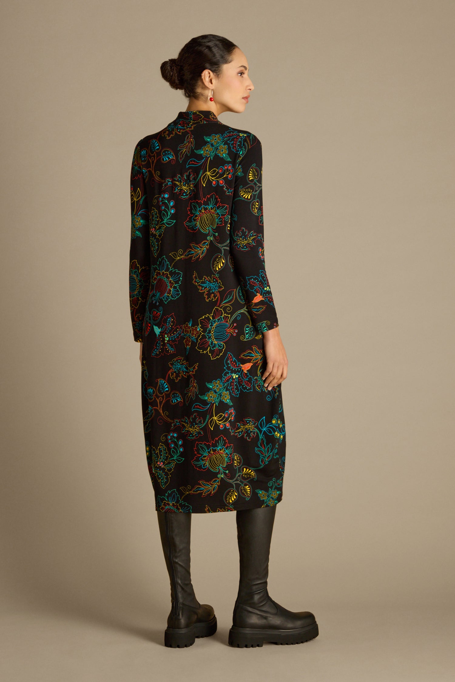 A woman with her hair styled in a bun stands against a plain beige background, wearing the Bright Dotted Floral Jersey Dress featuring a V-neckline, paired with black knee-high boots. She is facing away from the camera.